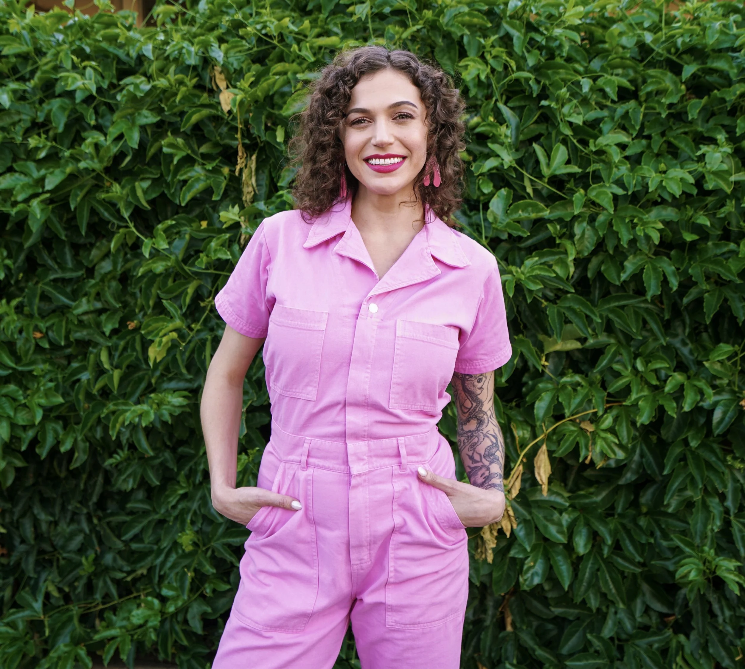 Cady S. Moore, AASECT-certified sex educator, standing and smiling in a full-length pink jumpsuit in front of a lush green leafy wall