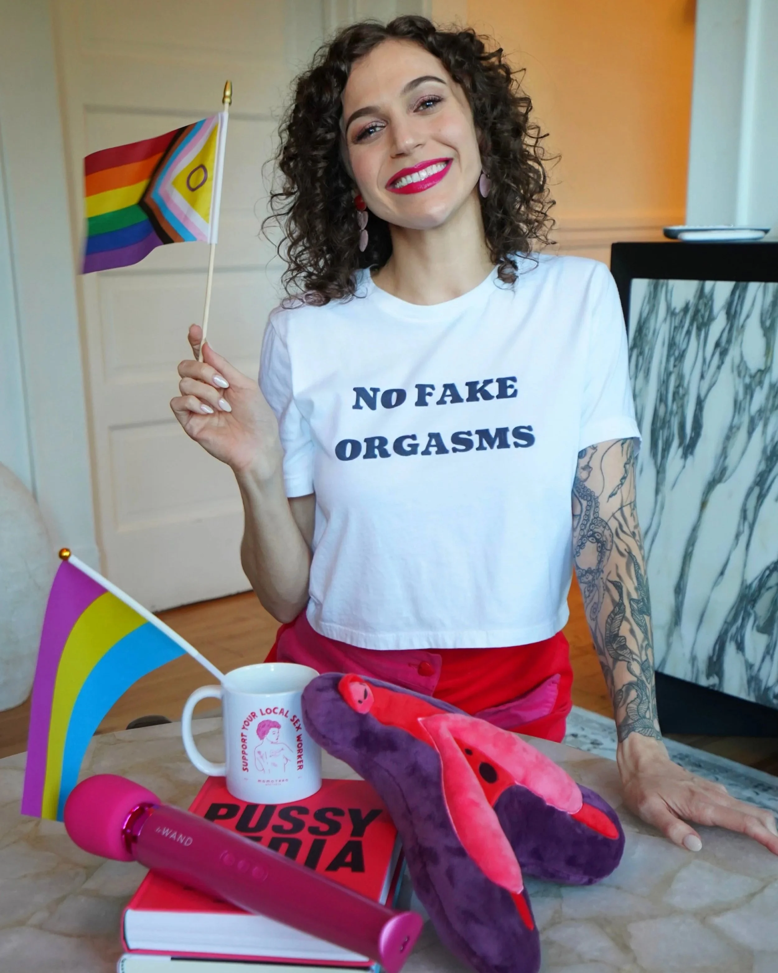 Cady S. Moore wearing a No Fake Orgasms t-shirt holding an intersex-inclusive pride flag, seated with sex education books, a Le Wand vibrator, and a Support Your Local Sex Worker mug