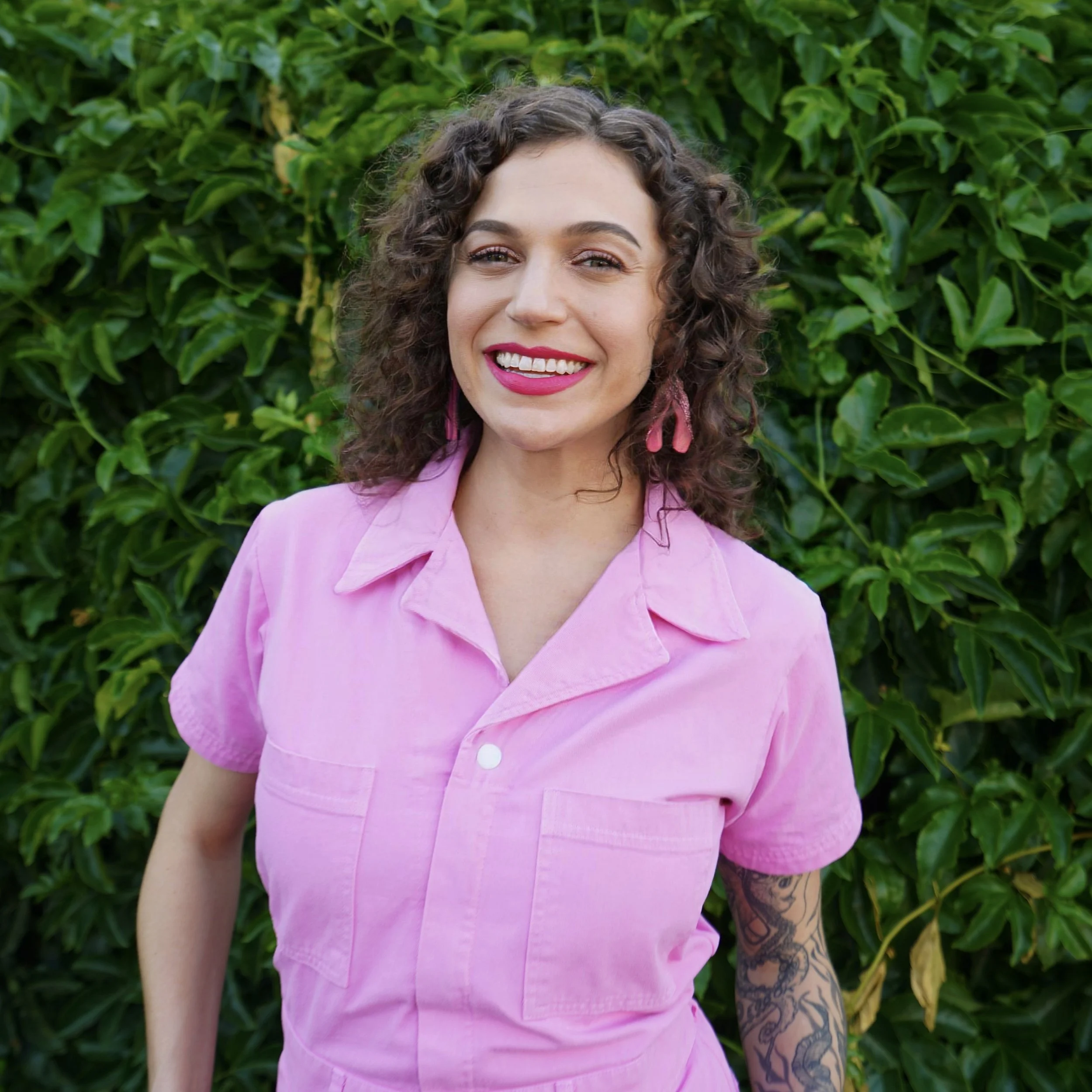 Cady S. Moore, AASECT-certified sexuality educator and coach, smiling in a pink jumpsuit in front of a green leafy wall in Sacramento
