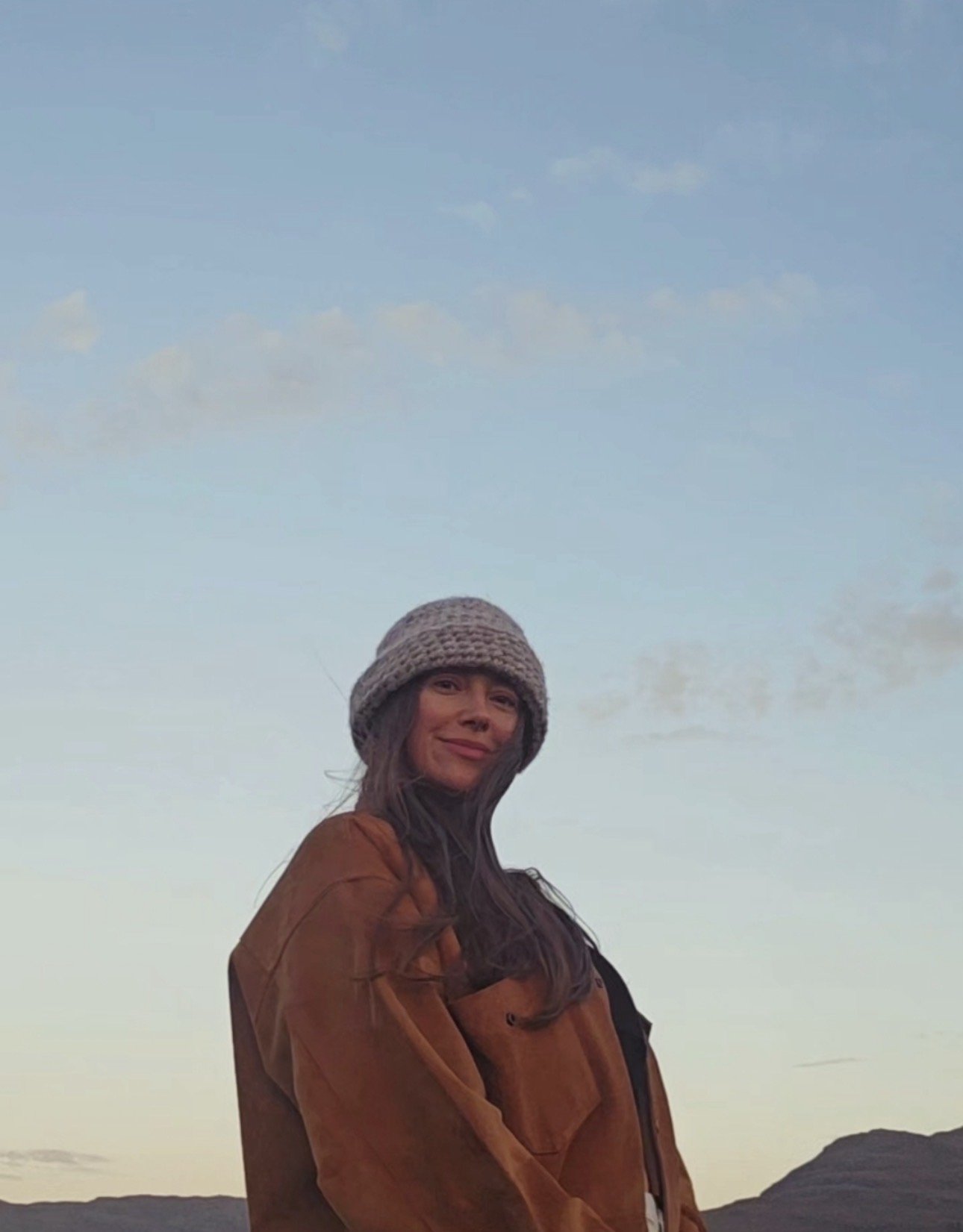 A woman with long dark hair, wearing a gray knit hat and a brown jacket, stands outdoors at sunset or dusk with a landscape and sky in the background.