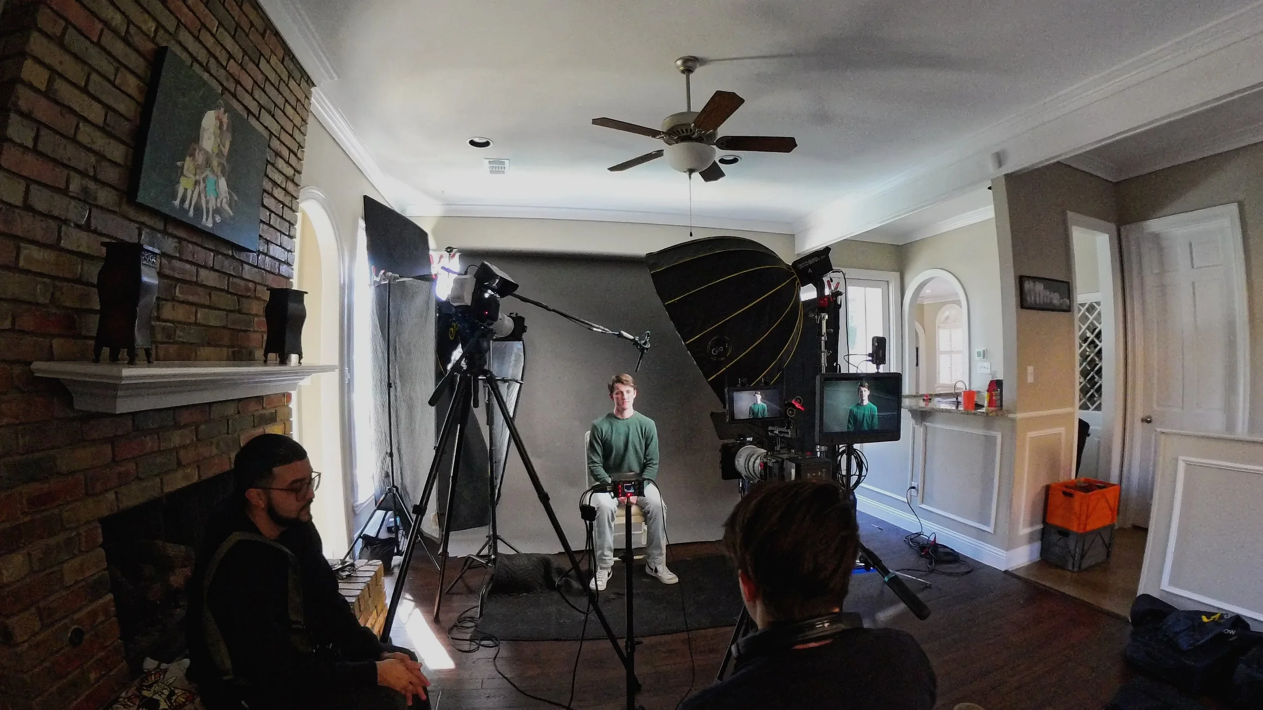Indoor video shoot setup with a young man sitting in front of a gray backdrop, surrounded by film equipment including cameras, lights, and monitors in a room with a brick wall and ceiling fan.