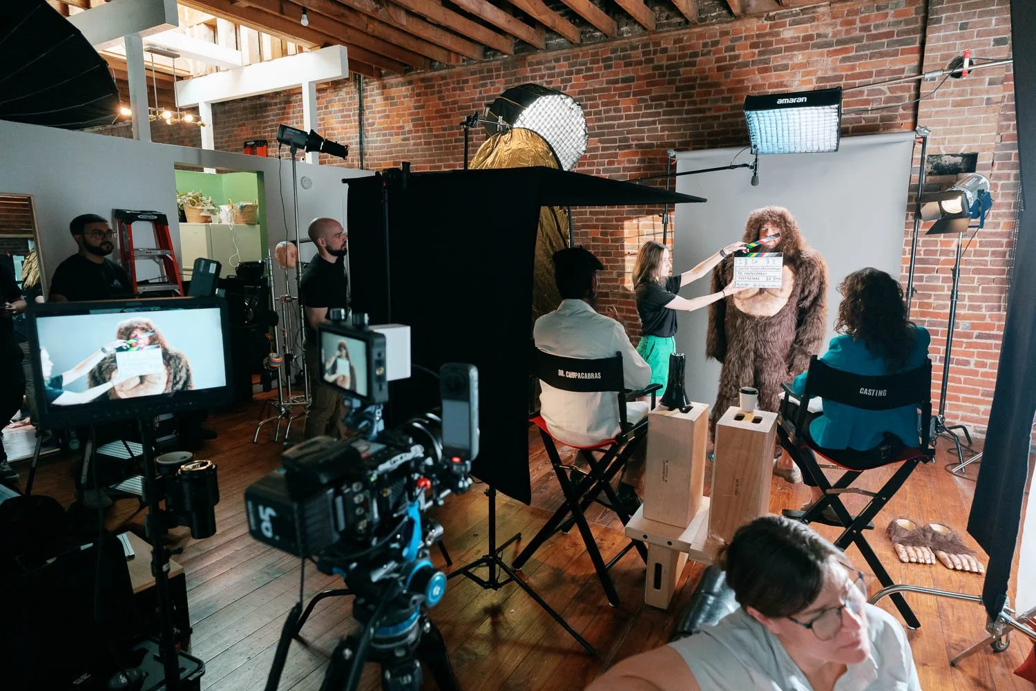Film crew on set with actor dressed in a big furry costume for a photo shoot or film scene, with equipment, crew members, and a brick wall background.
