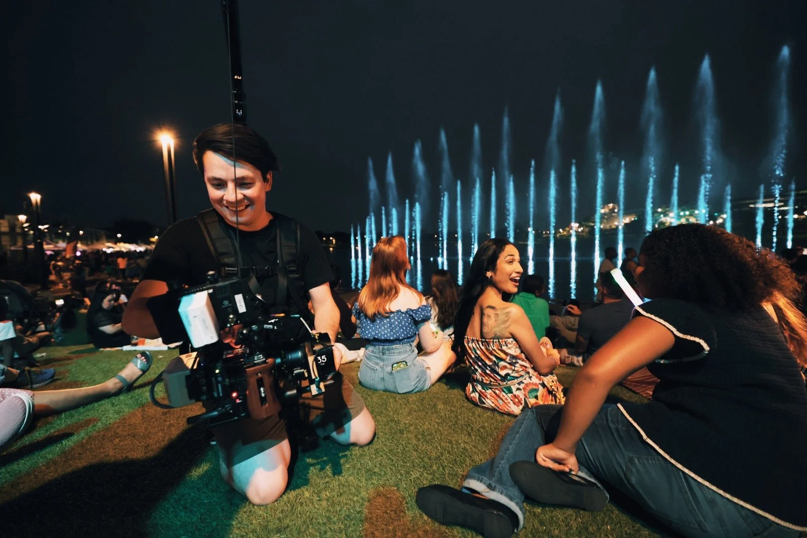 People sitting on grass at night watching a fountain light show with blue water jets.