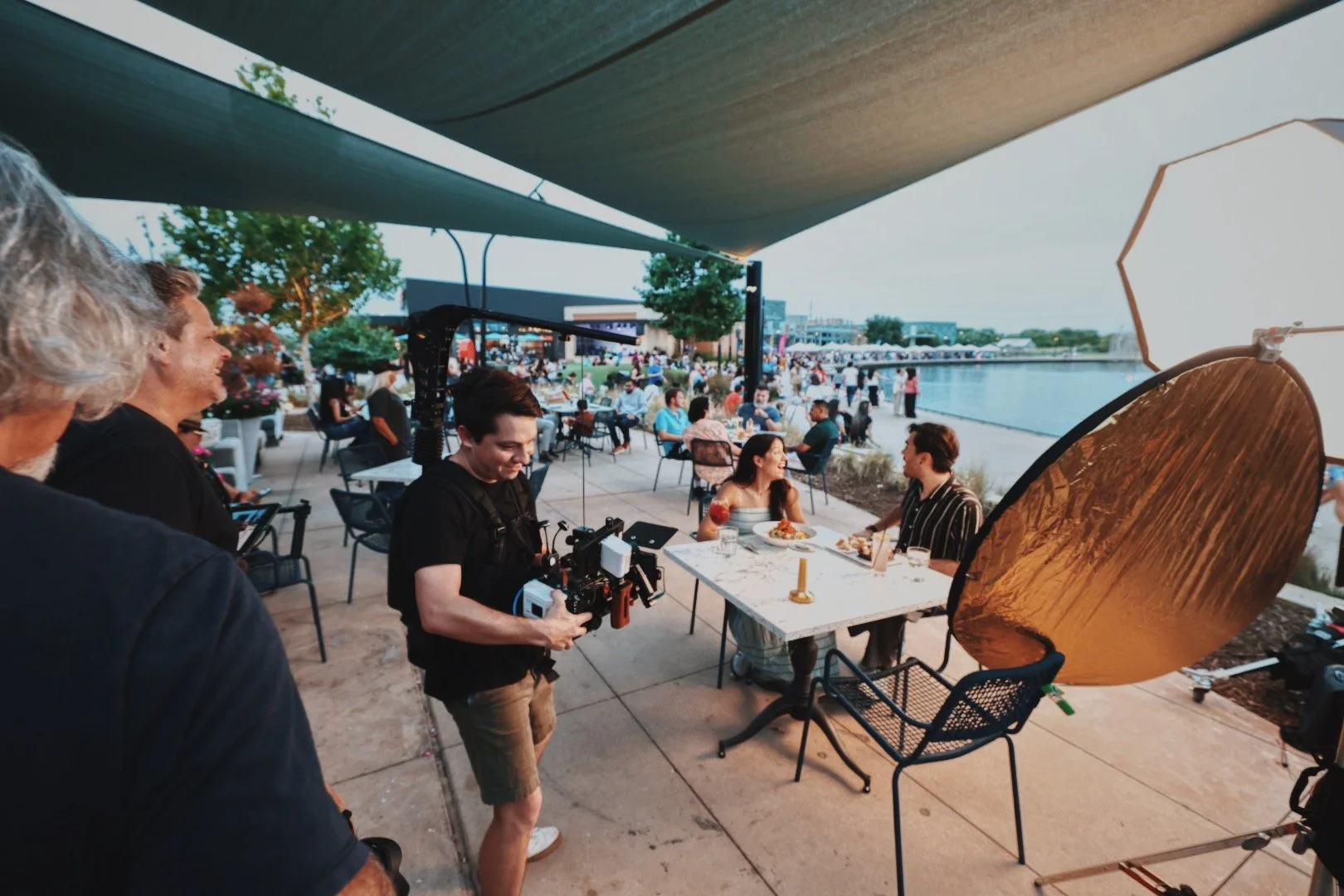 Filming crew setting up a scene with two women at an outdoor waterfront restaurant during the evening.