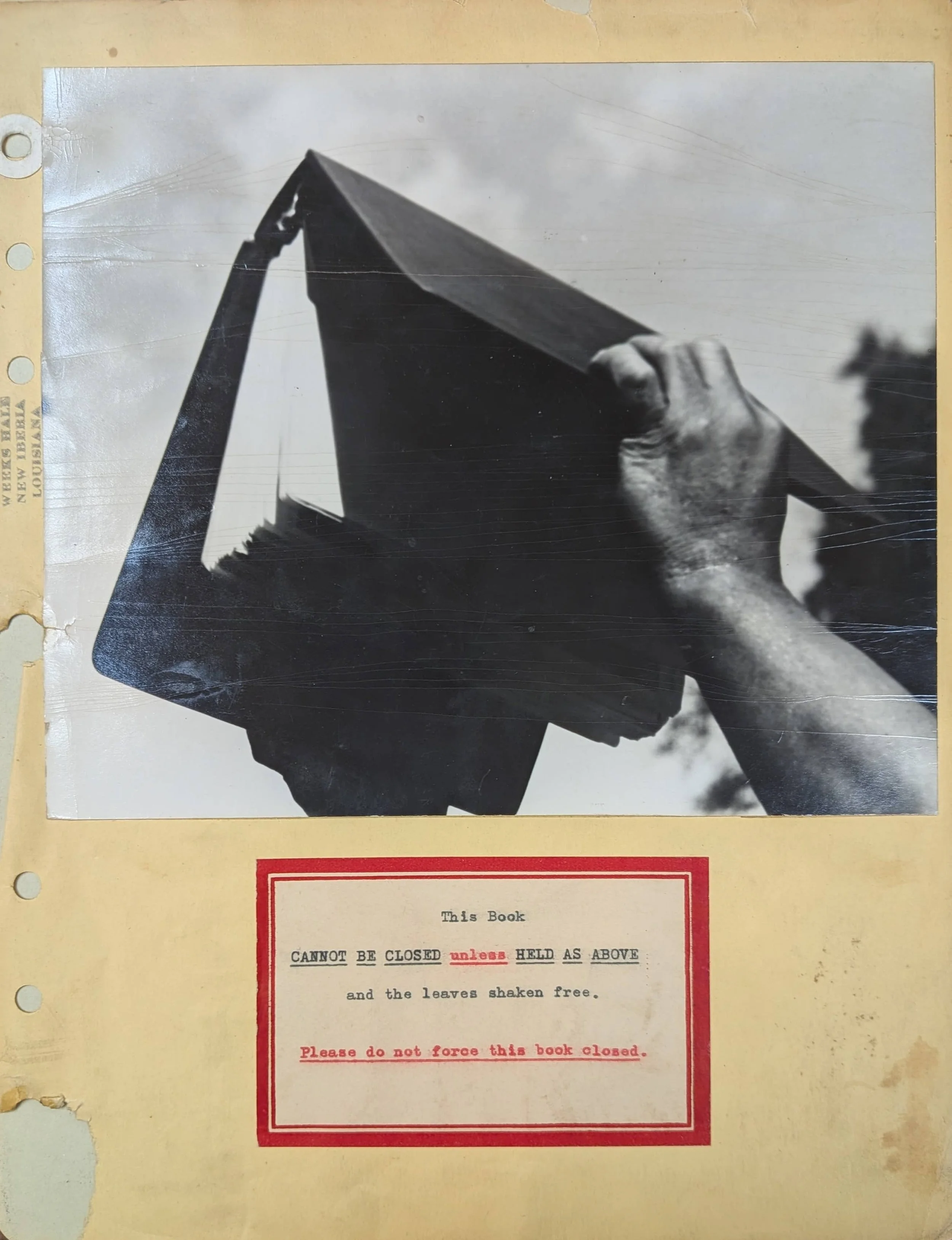 Black-and-white photo of a hand holding a book upright so its pages hang downward; below it, a typed label reads: “This Book CANNOT BE CLOSED unless HELD AS ABOVE and the leaves shaken free. Please do not force this book closed.”