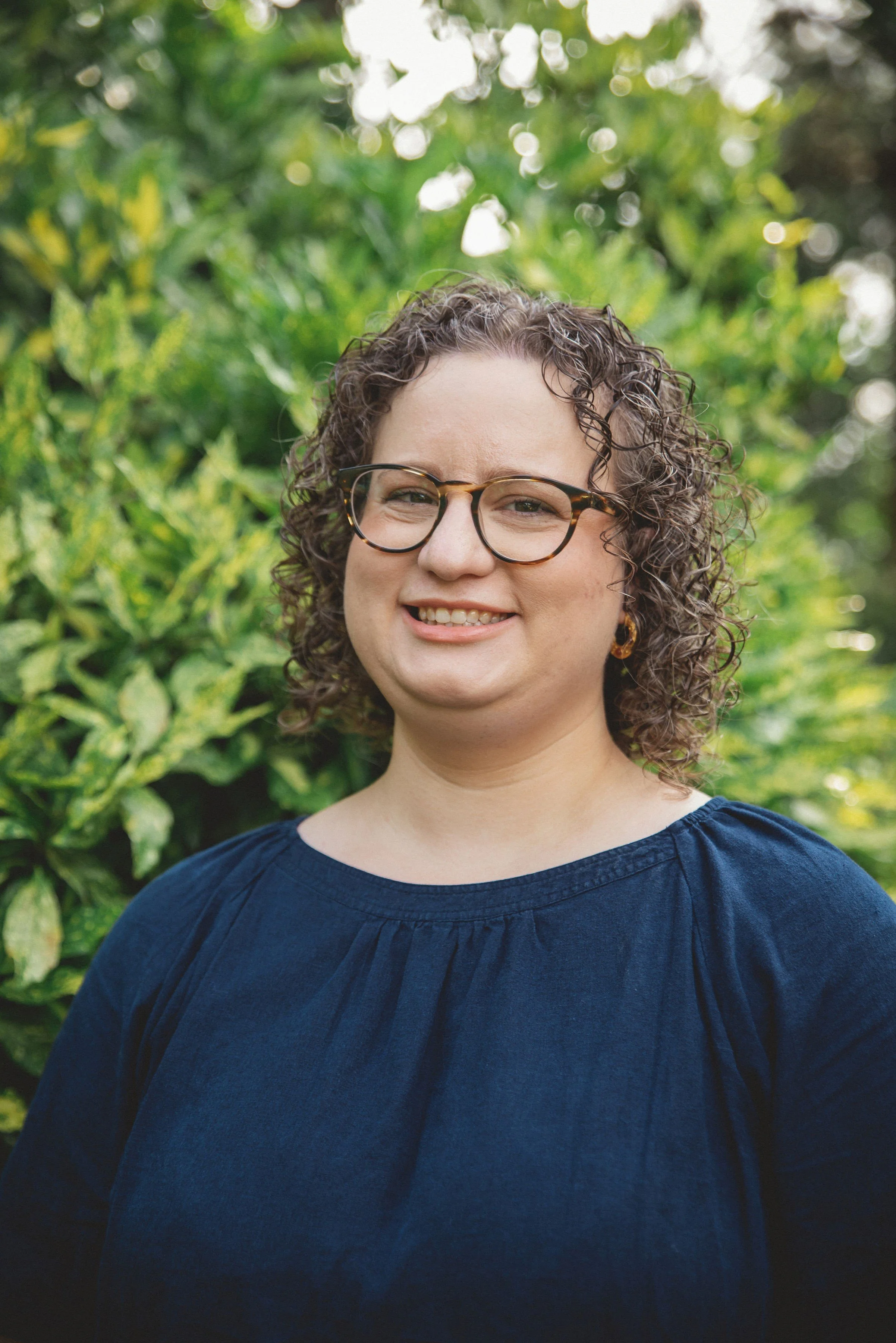 Portrait photograph of Sarah Duggan, a white woman with curly brown hair and tortoiseshell glasses, wearing a navy blue top, photographed outdoors.