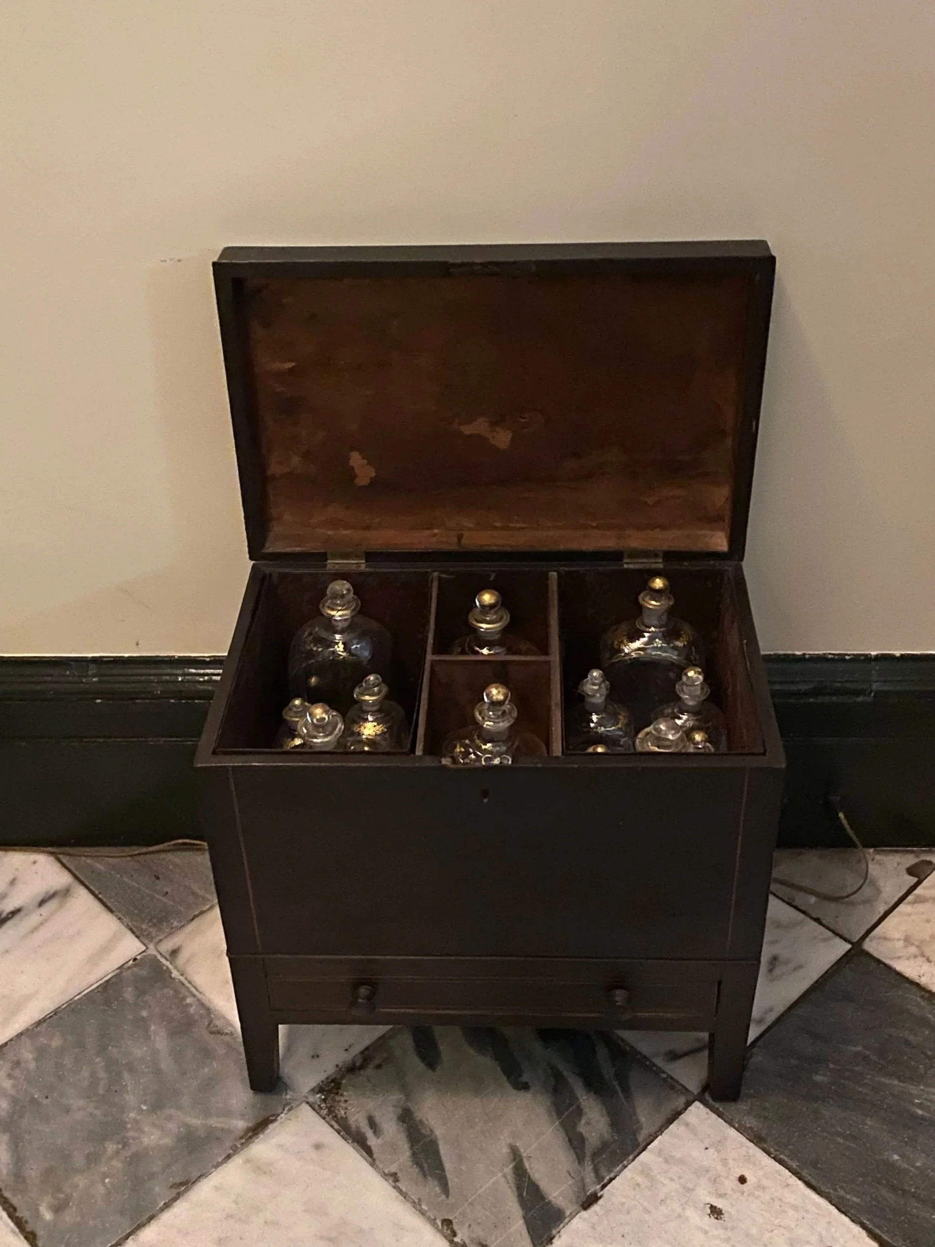A dark wooden cabinet with its door open, revealing several decorative glass decanters and bottles stored on interior shelves. The cabinet stands against a light-colored wall beside a blue-and-white tiled floor.