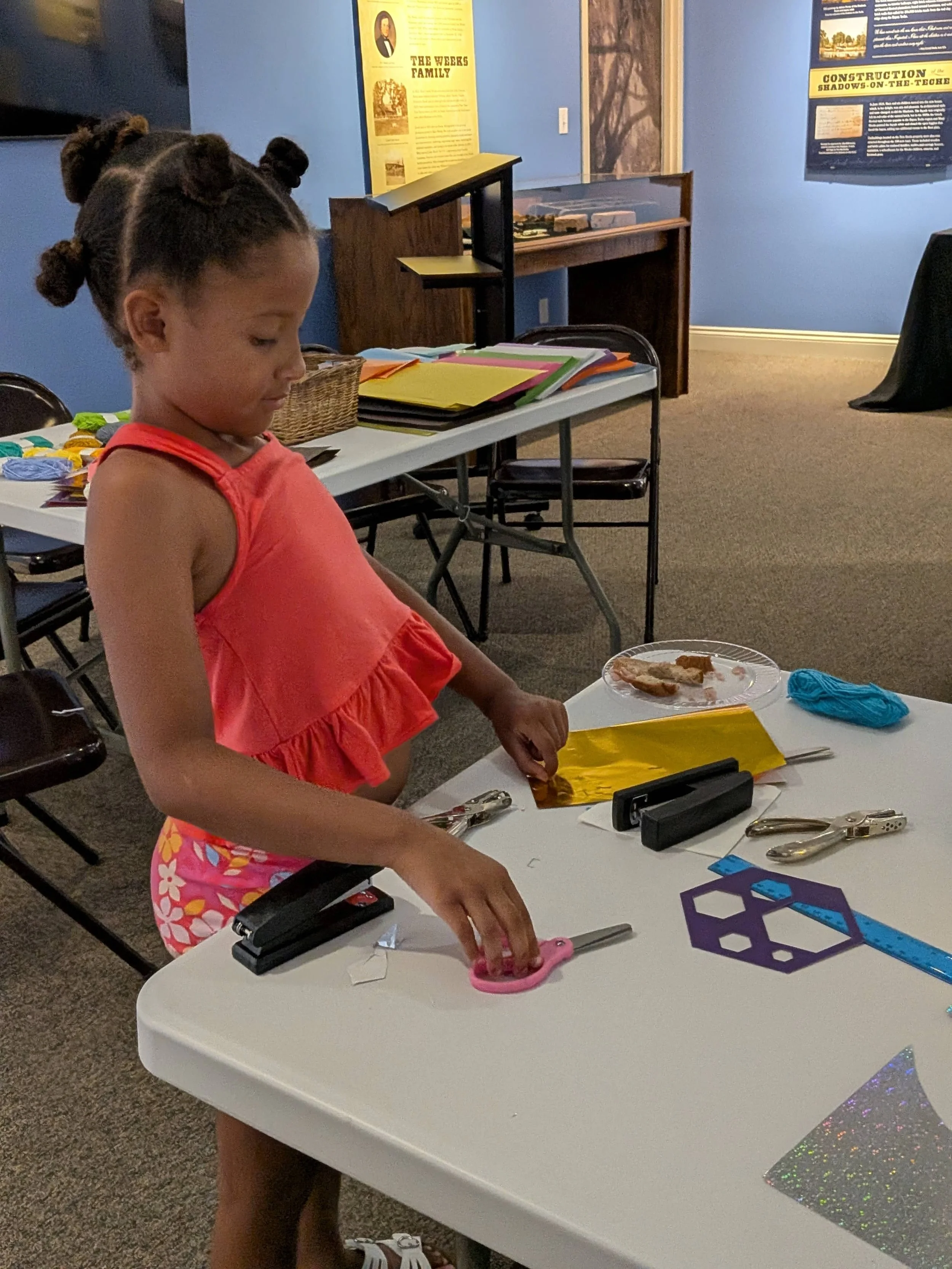 Girl with hair in twisted buns crafts a lantern using gold paper and scissors at a table in a museum classroom.