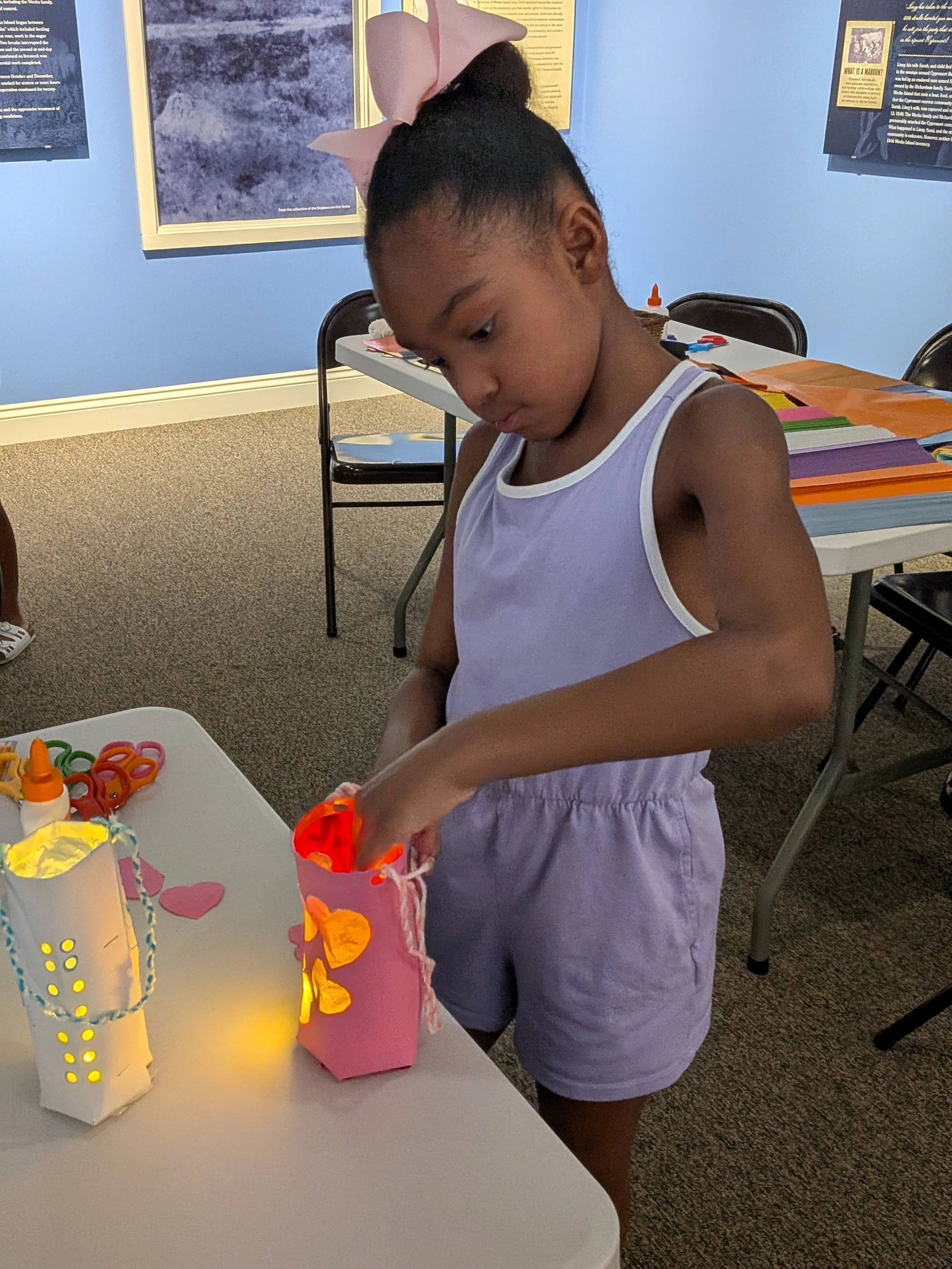 Girl with a pink bow reaches into her glowing pink paper lantern, another lit lantern beside her on the table.
