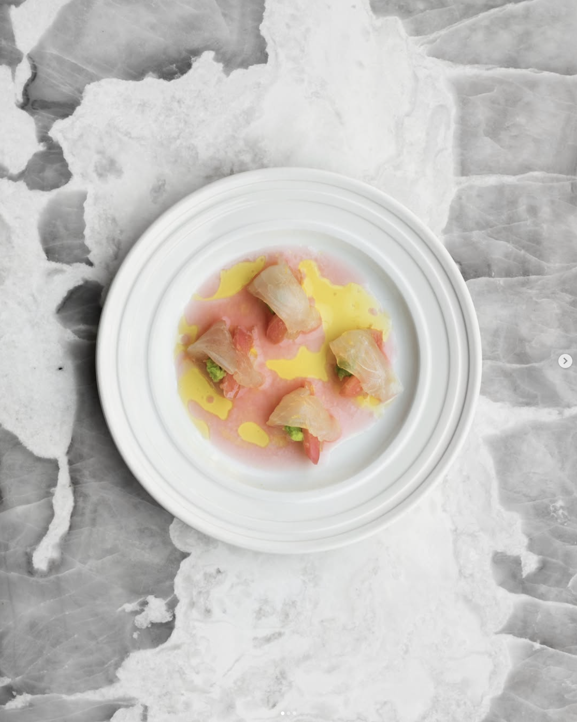 An overhead shot of a crudo seafood dish artfully plated on a white plate, on top of a white and gray marble surface