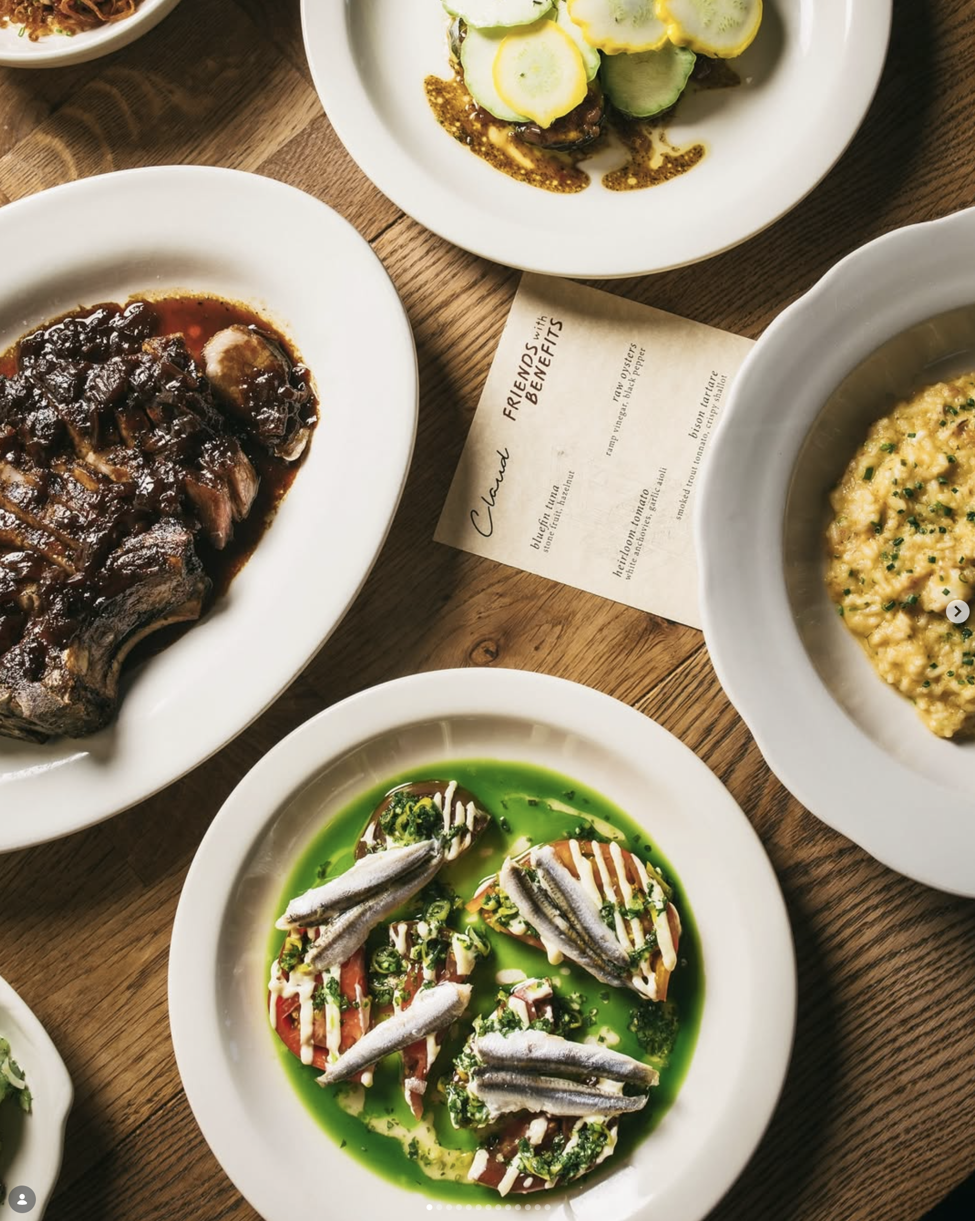 An overhead with multiple dishes on a table, including a sliced steak, and an anchovy and tomato dish. There is a menu in the center