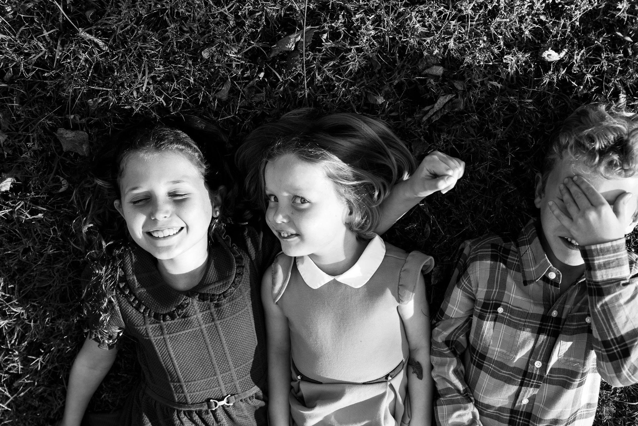 Three children are laying on their backs in the grass. One child smiles and closes her eyes. The middle child gives the camera a mischievous look. The next child covers his eyes with one hand.