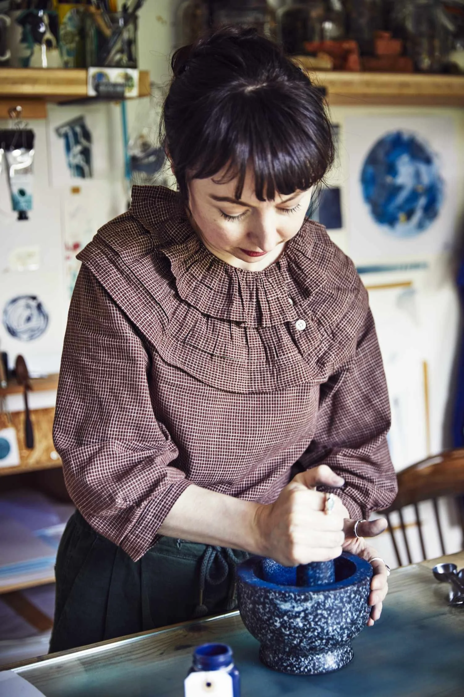 A brown haired woman wearing a checked ruffled collar shirt is grinding blue pigment in a pestle and mortar.