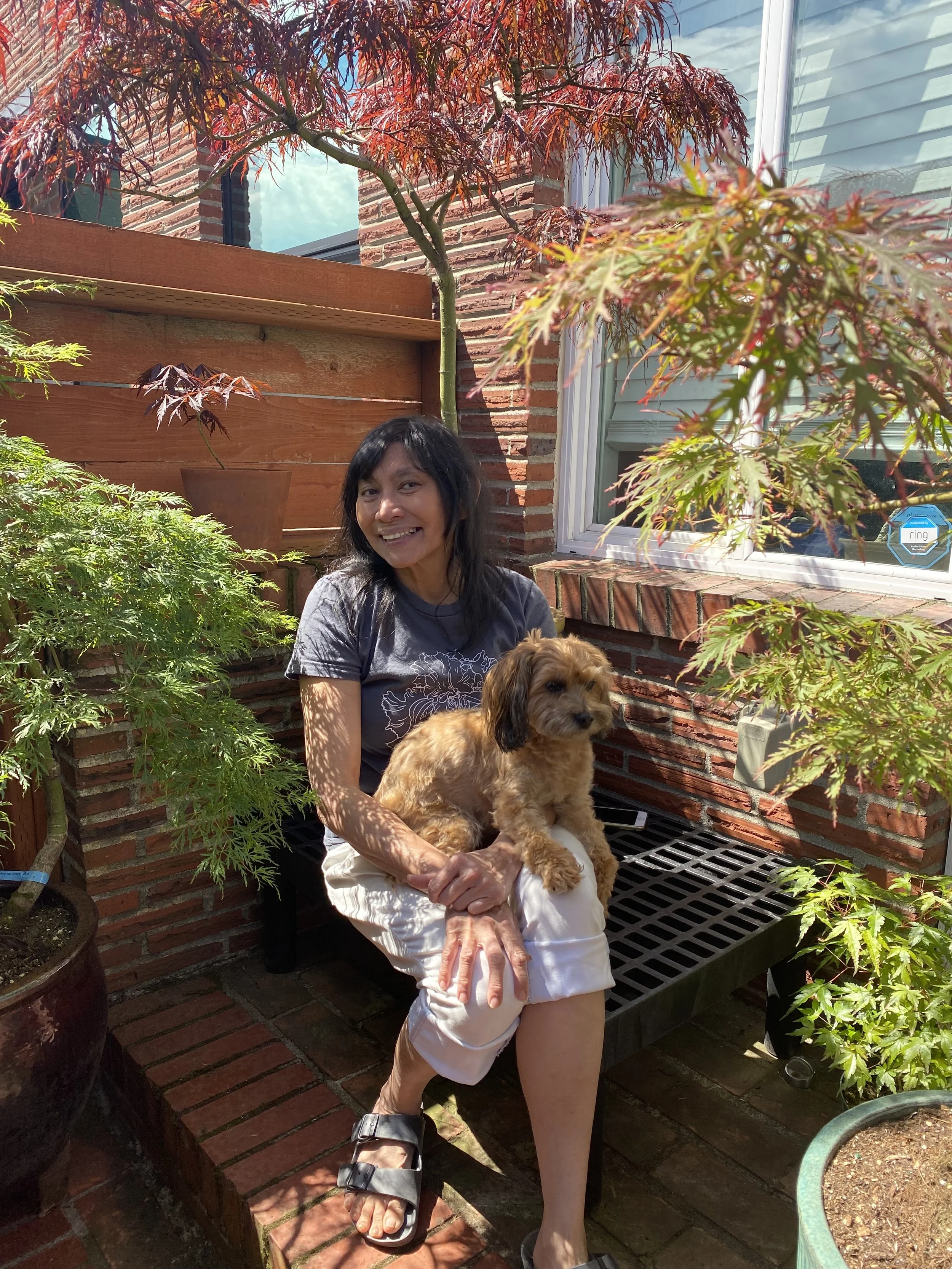 Cynthia Alexander, the sole owner, operator, and designer of Flora Tacoma sitting outdoors under a red and green Japanese maple tree, holding her small poodle-mix, Rosie, in front of her little brick home studio in the heart of Hilltop, Tacoma.