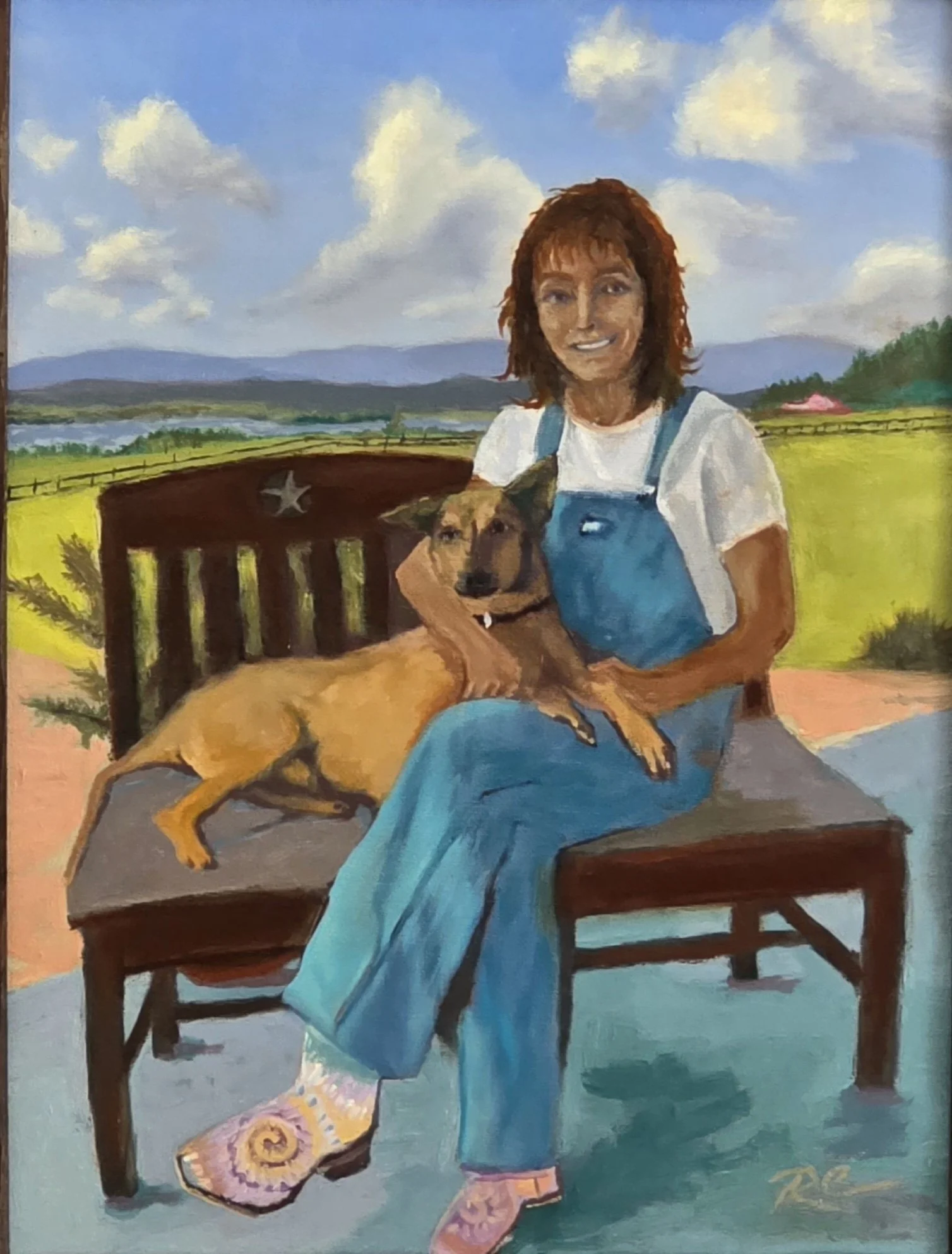 A woman with reddish-brown hair sitting on a wooden bench outdoor with a brown dog. The woman is wearing a white t-shirt and blue overalls, and the dog is lying lightly on its side, resting its head on the woman's arm. The background features a scenic landscape with green fields, a river, hills, and a partly cloudy sky.