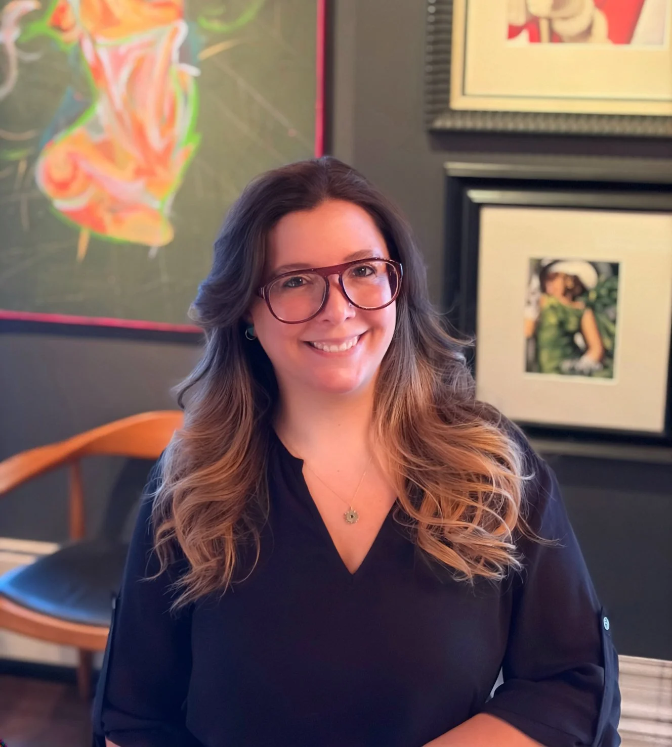 Smiling woman with long wavy brown hair wearing glasses and a black top, seated indoors with framed artwork on the wall behind her.