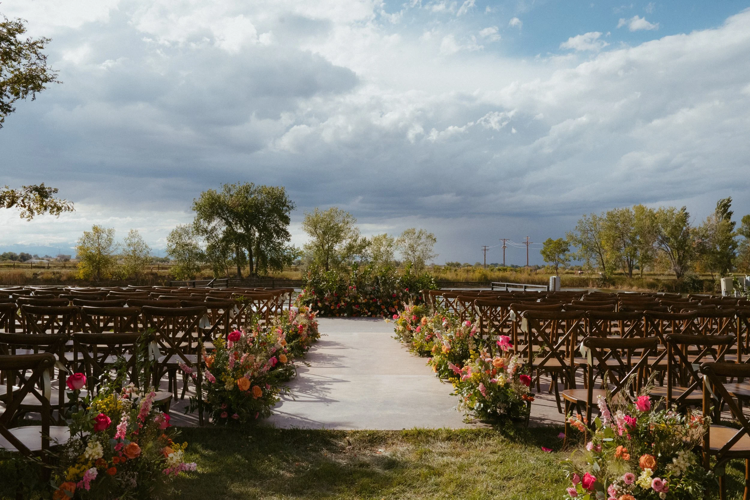 Outdoor wedding setup with floral arrangements along an aisle, chairs on either side, and a cloudy sky overhead.