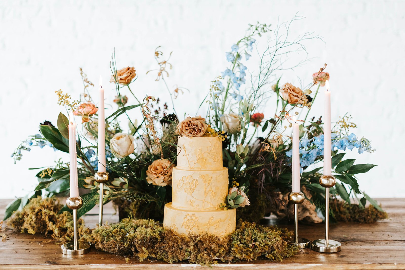 Elegant three-tier yellow wedding cake decorated with flowers, surrounded by lush greenery, tall pink candles in silver holders, and dried moss on a wooden table.
