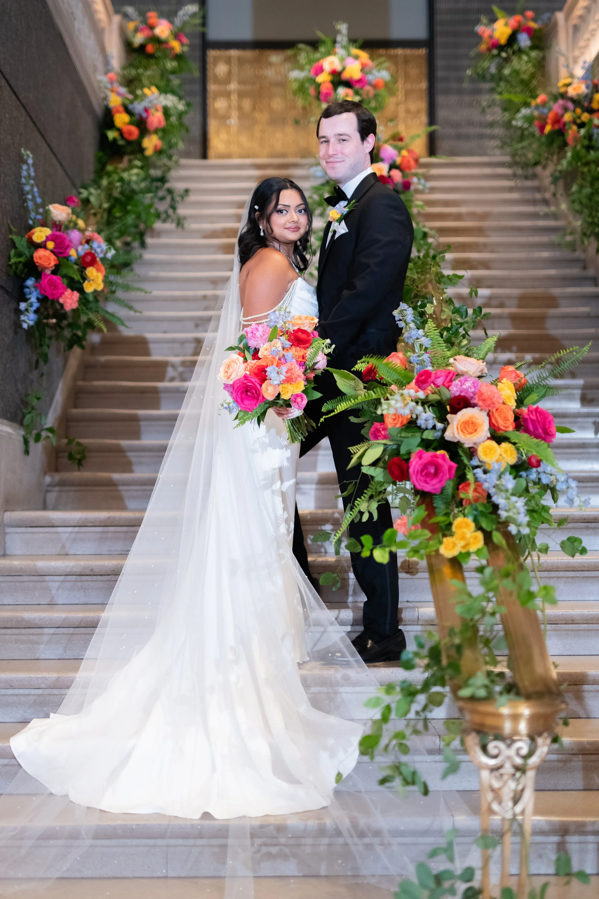 A bride and groom standing on stairs during their wedding, surrounded by colorful floral arrangements.
