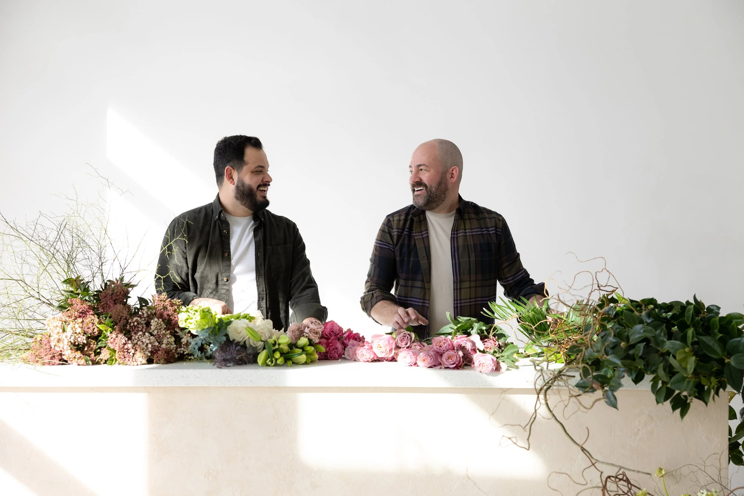 Two men smiling and talking at a table with various flowers and greenery in a bright, minimalistic room.
