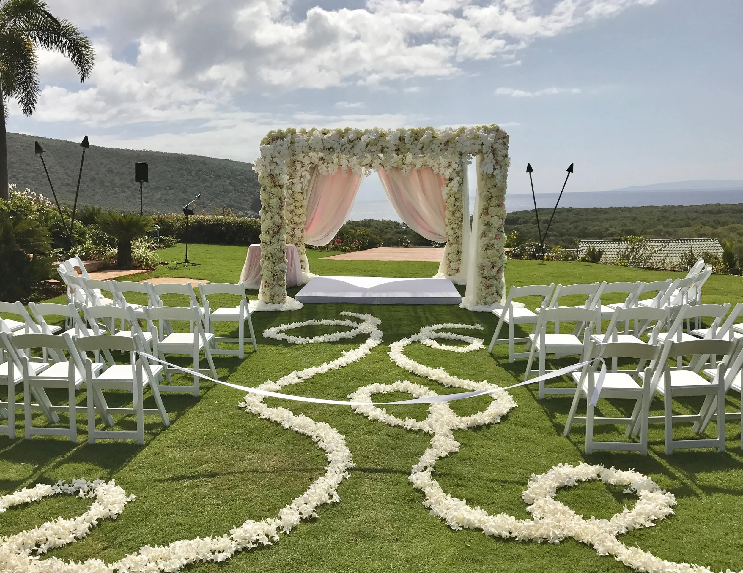 Outdoor wedding ceremony setup with white chairs, a flower arch, and floral-designed paths on a grassy area with scenery of hills, trees, and the ocean in the background.
