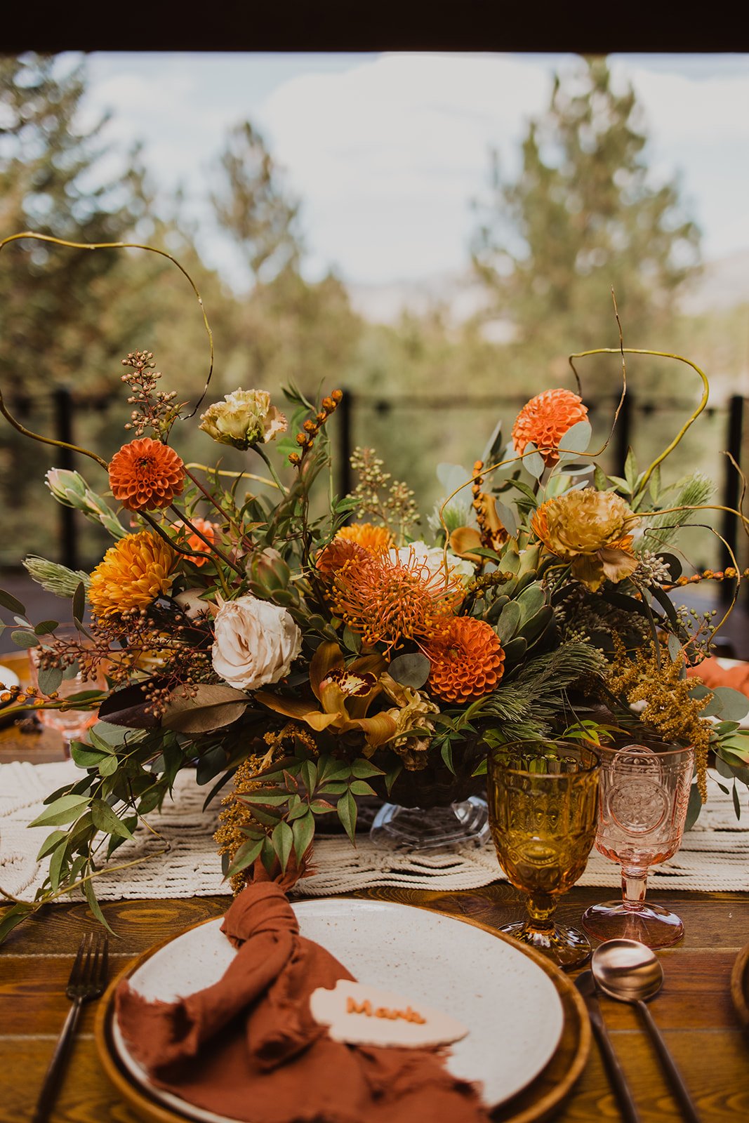 A table setting with a large floral centerpiece featuring orange, cream, and yellow flowers, surrounded by green foliage and twisting twigs. The table has a white placemat, gold-rimmed white plate with a rust-colored cloth napkin, a small white heart