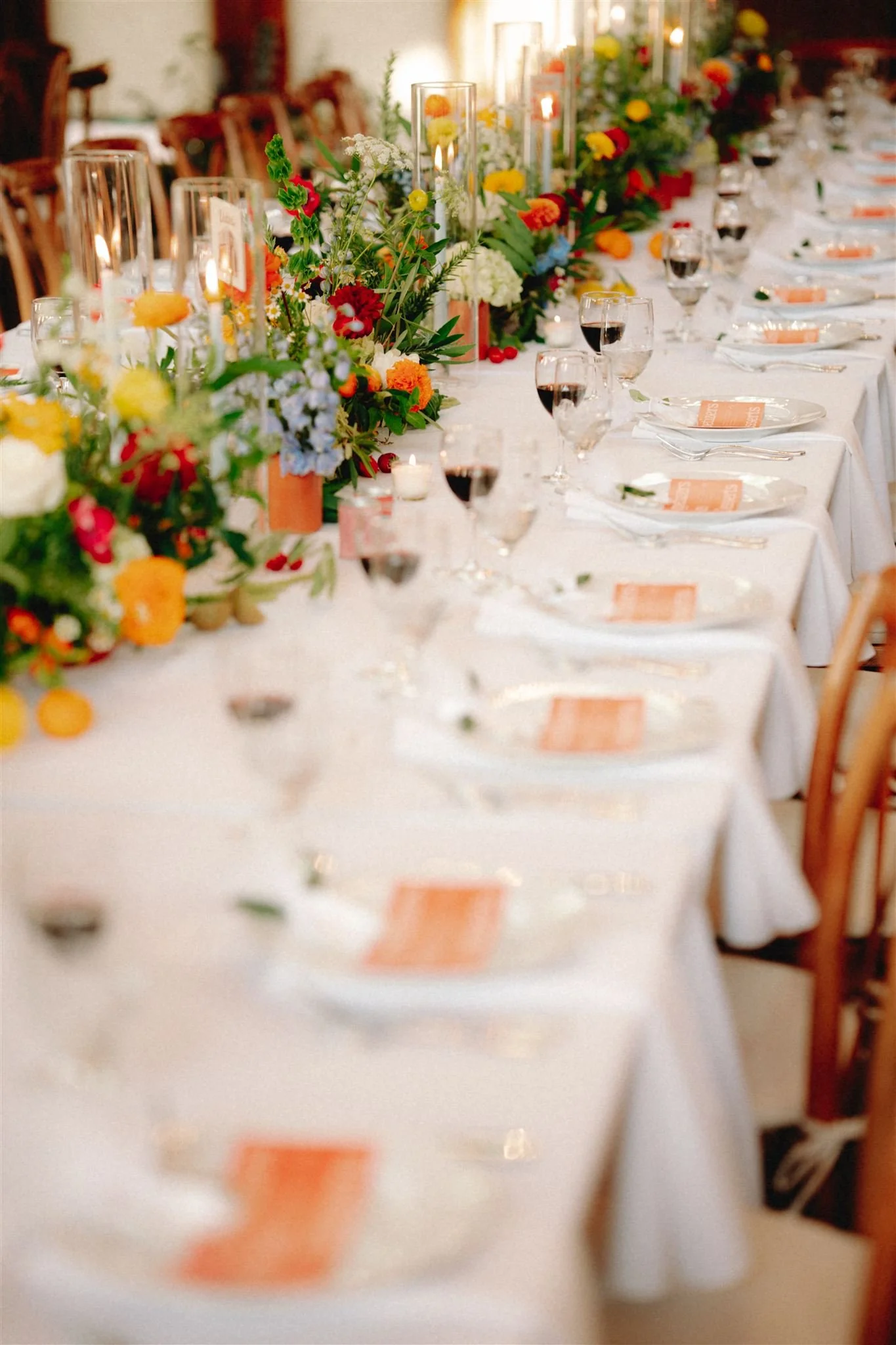 A long banquet table decorated with colorful flowers, candles, glassware, and place settings, set for a formal event or celebration.
