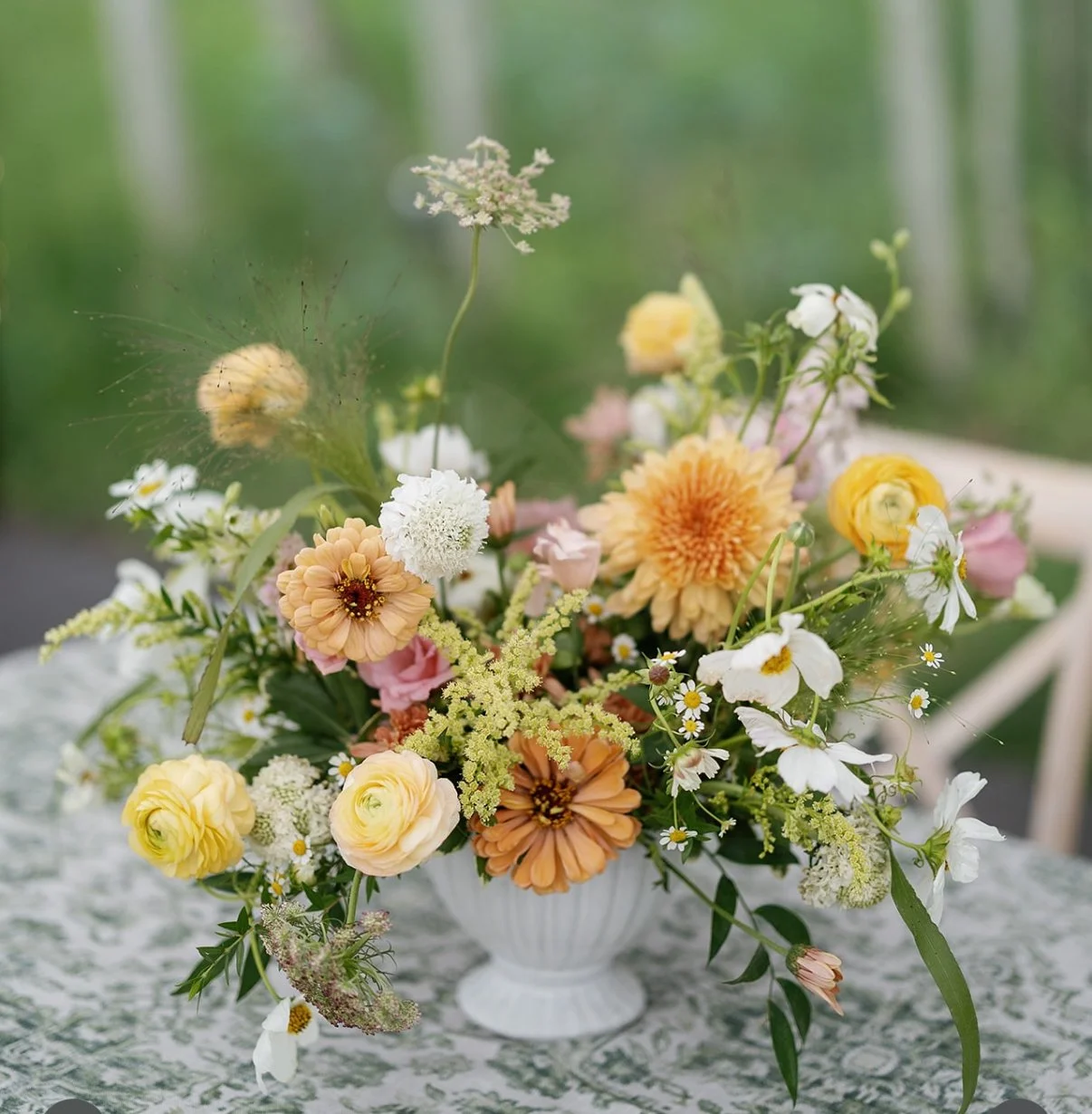 A floral arrangement in a white vase with various pastel-colored flowers on a patterned tablecloth, with a blurred green background.