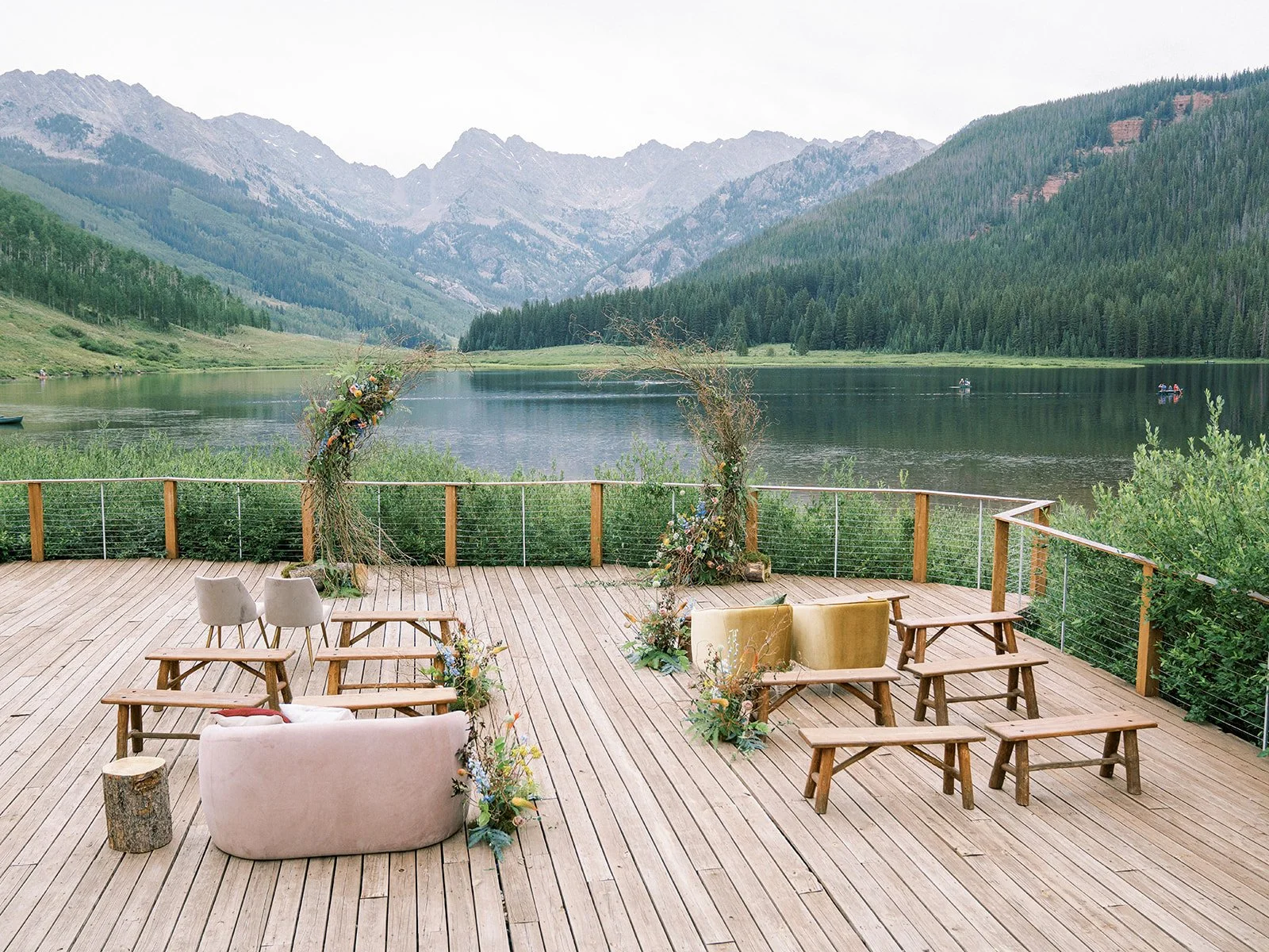 Outdoor wedding ceremony setup on a wooden deck overlooking a calm lake surrounded by mountains and dense forest, with chairs arranged for guests and floral decorations around a wedding arch.