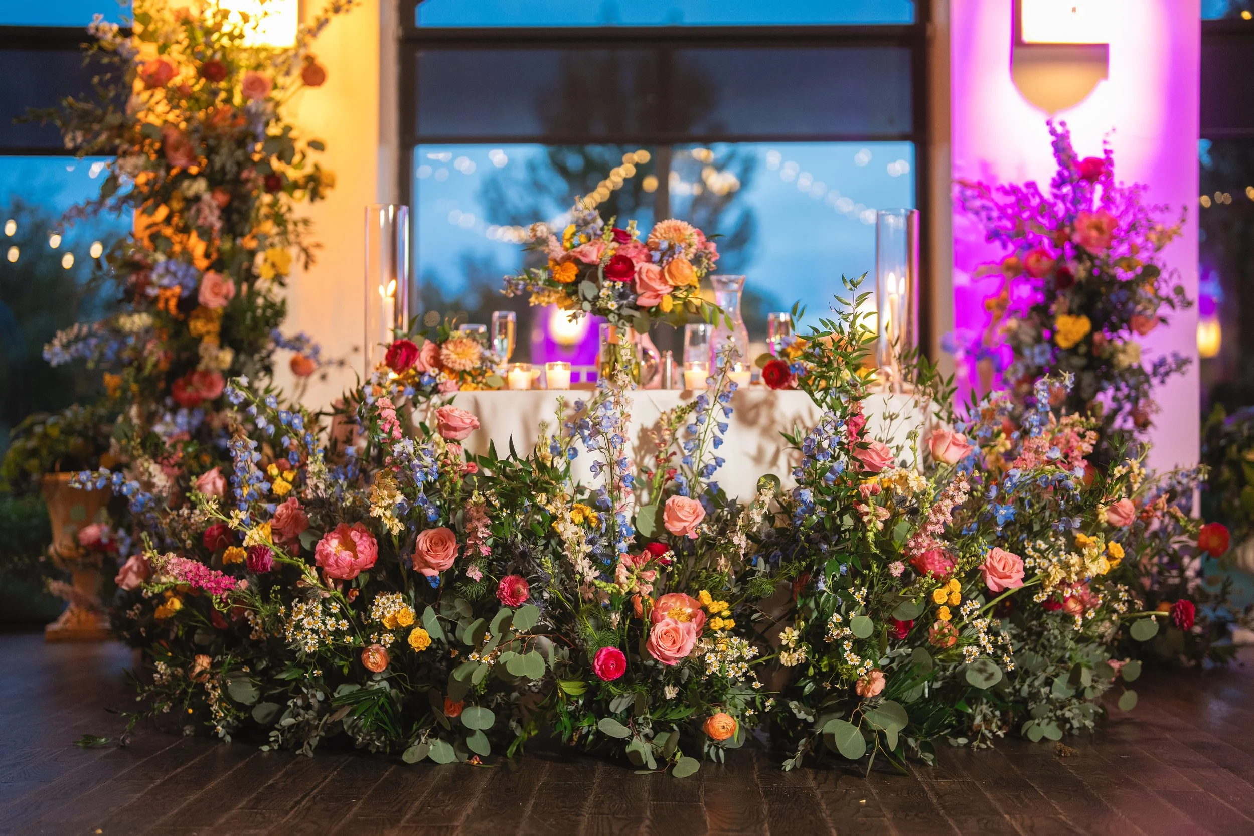 A decorated table with pink, yellow, and white flowers, surrounded by more flowers on the floor, and lit candles in glass holders, with large windows and purple and yellow lighting in the background.