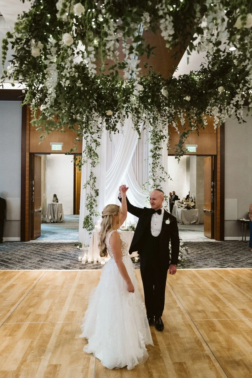 A bride and groom dancing at their wedding reception under a floral ceiling with hanging greenery and white flowers, with soft lighting and elegant decor.