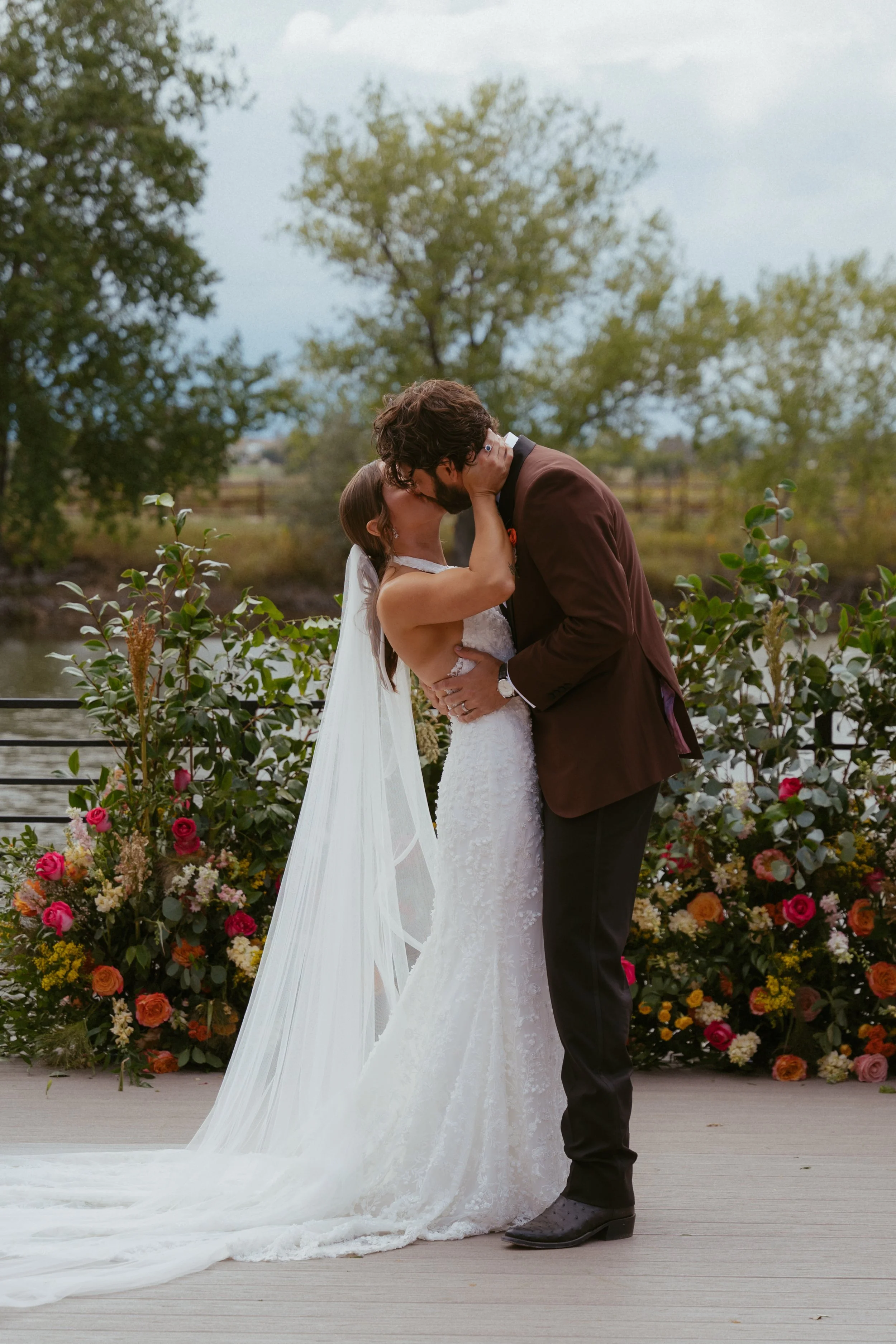 Bride and groom kissing outdoors with floral arrangements and trees in the background.