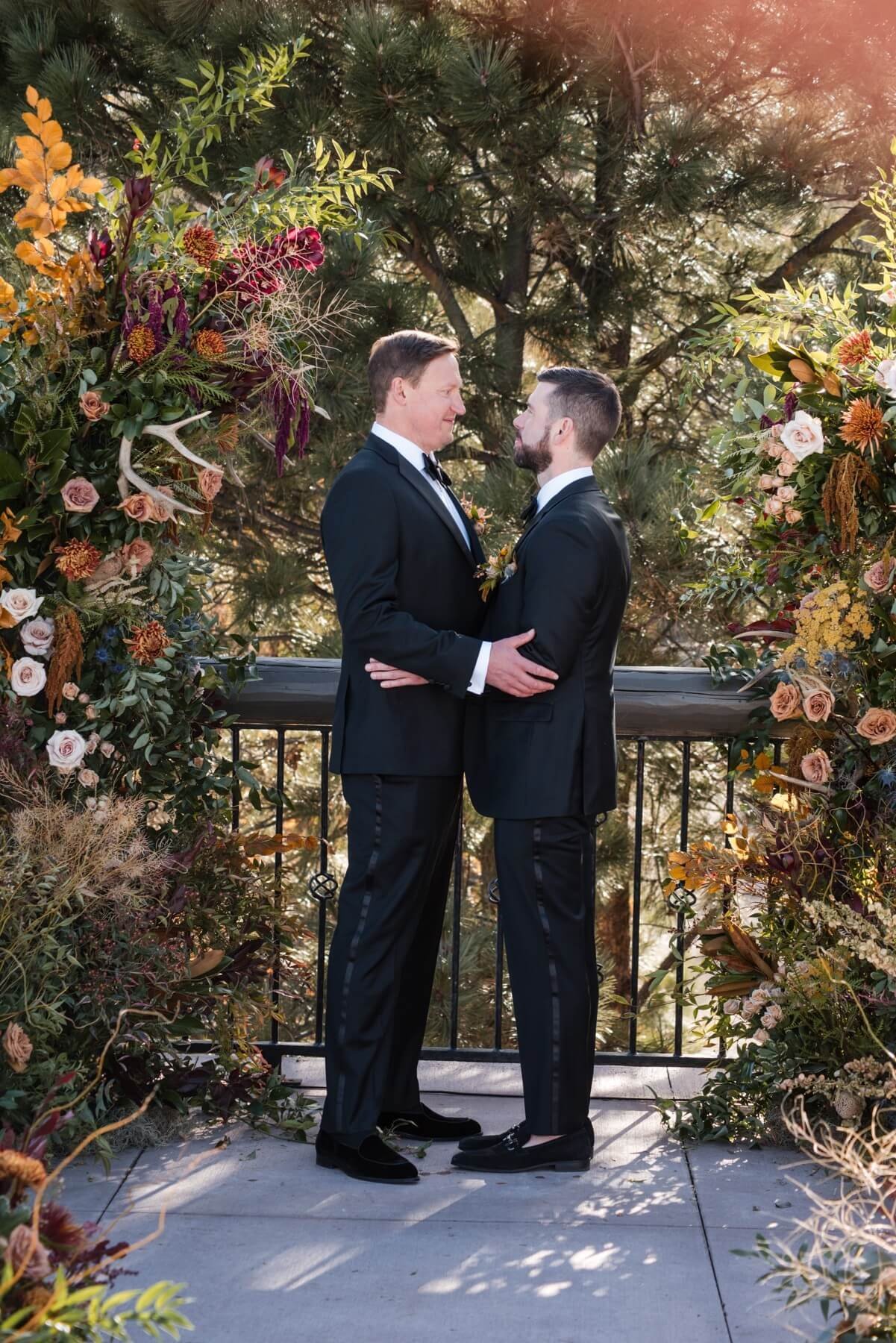 Two men in tuxedos holding hands and facing each other on a balcony framed by colorful floral arrangements, with trees in the background during daylight.