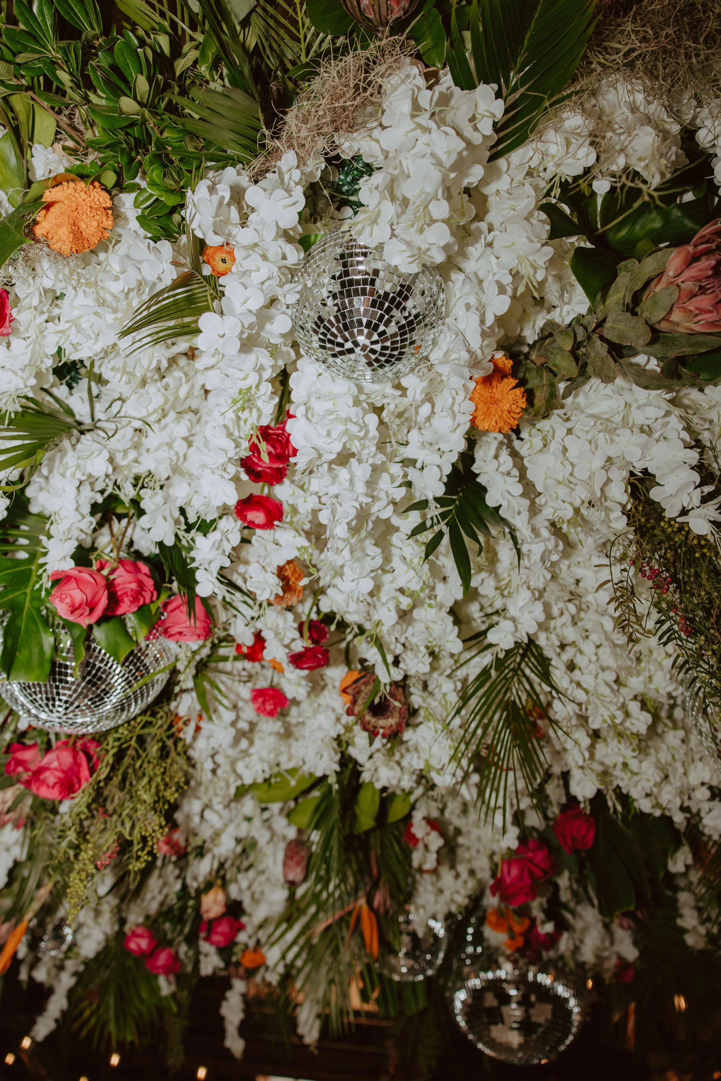 Close-up of a lush floral arrangement with white flowers, including hydrangeas and orchids, accented with colorful roses and marigolds, decorated with silver disco balls.