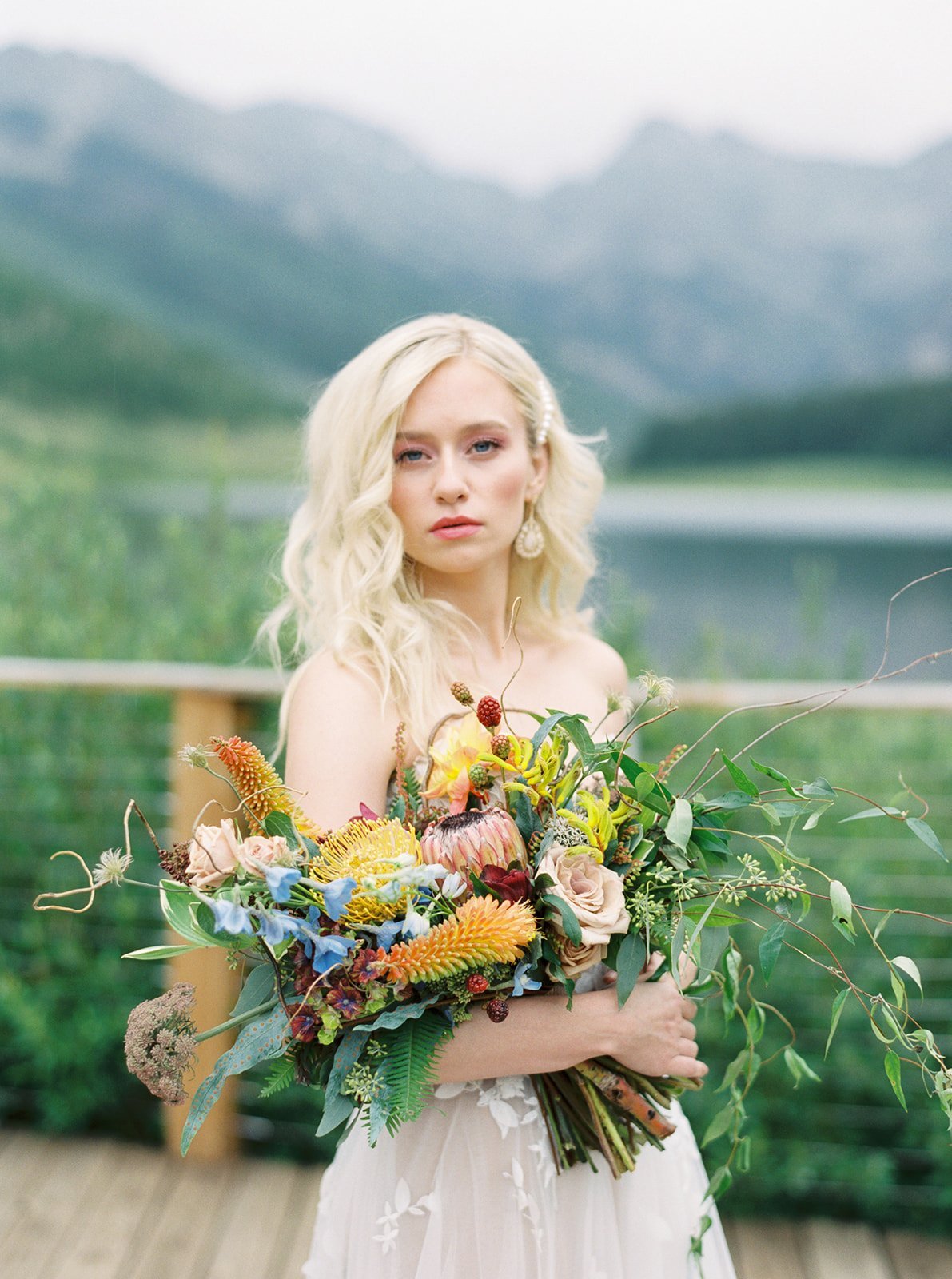 A woman with blonde hair holding a large bouquet of colorful flowers outside with a scenic mountain and lake background.