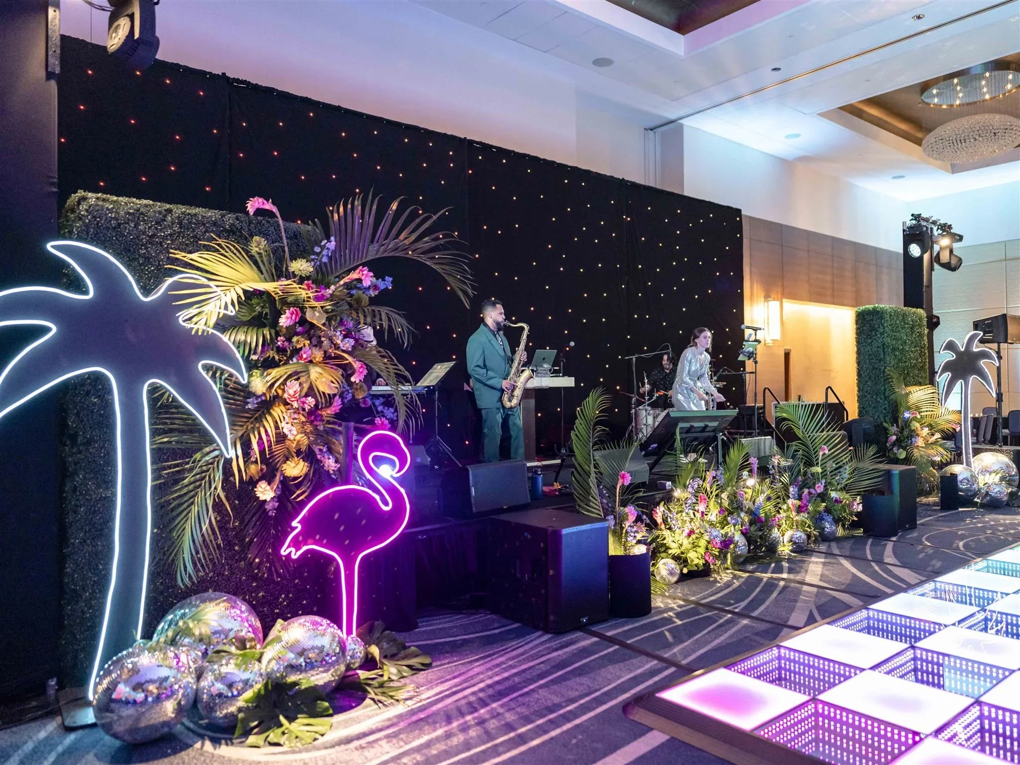 Indoor tropical-themed event space with neon flamingo and palm tree signs, decorated with flowers and greenery. Live band performing on stage with purple and yellow lighting.