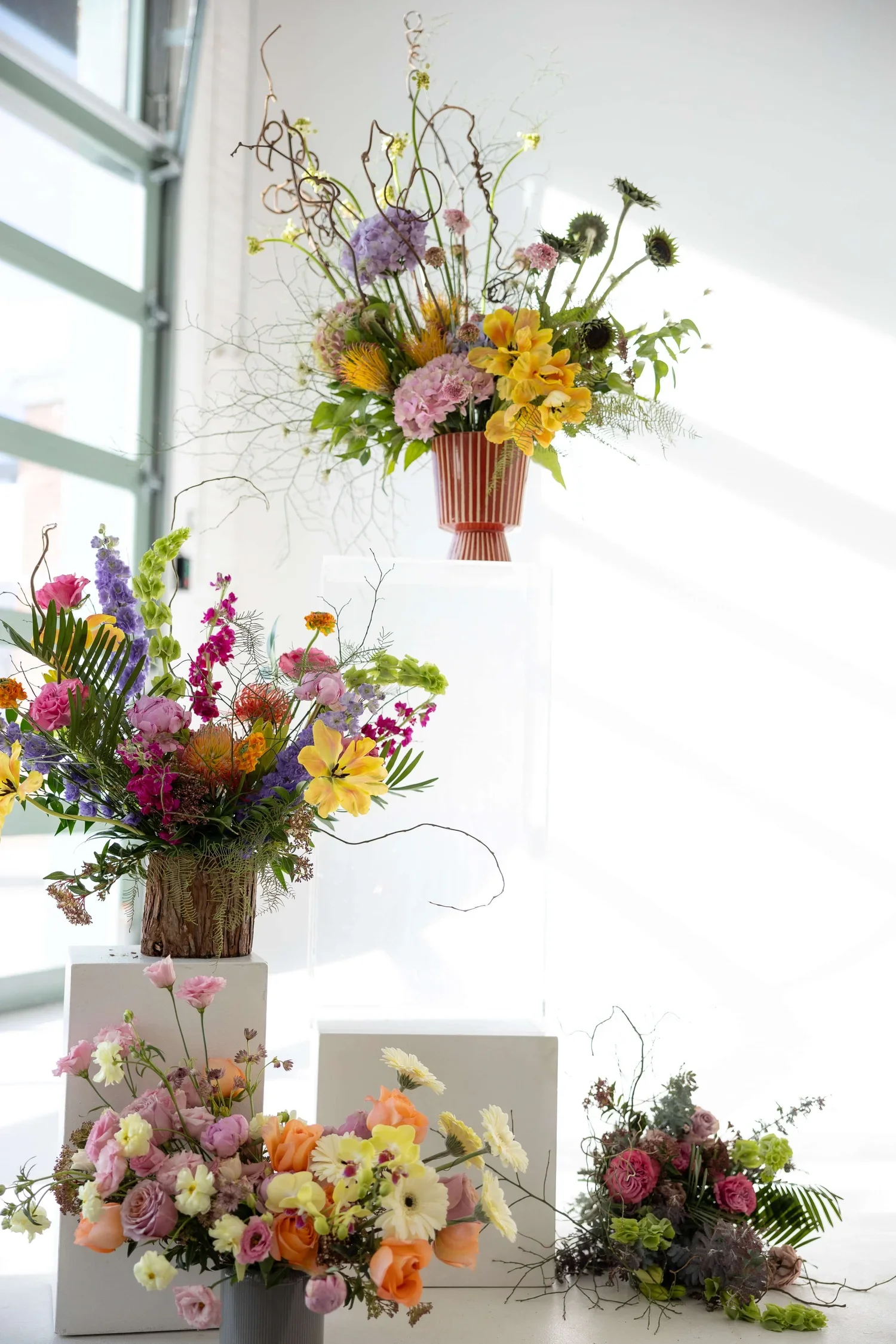 Arrangement of various colorful flower bouquets in vases on display, with some on white pedestals and others directly on the surface, against a light background near a window.
