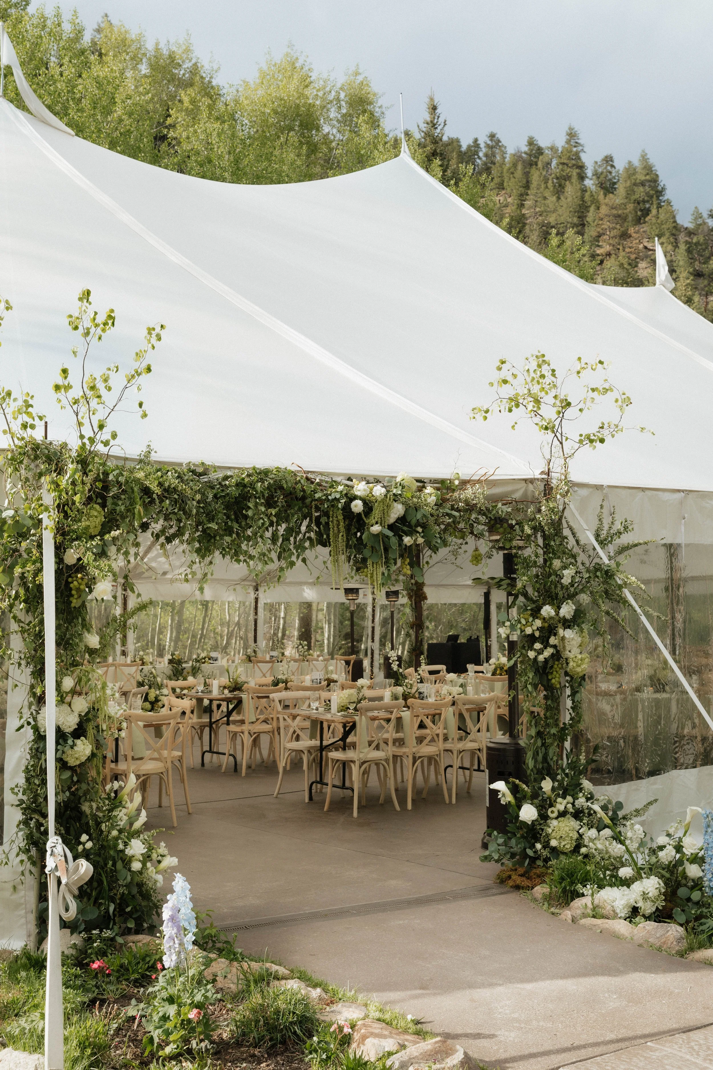 Outdoor wedding reception setup with tables, chairs, and floral decorations under a white tent with greenery and trees in the background.