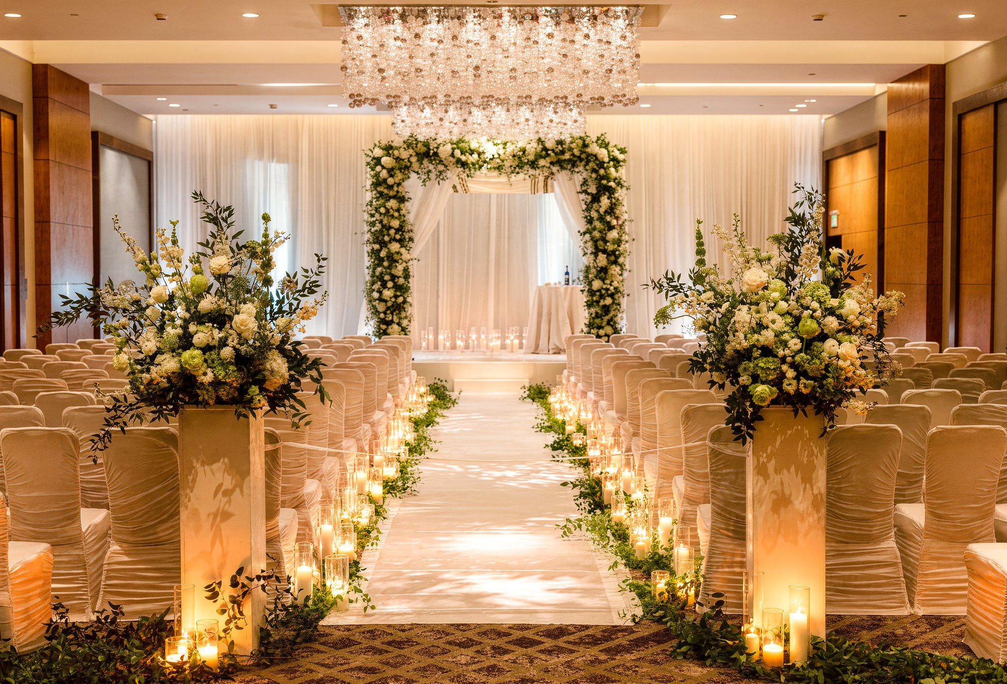 Wedding ceremony setup with white chairs, floral arrangements, candles, and an arch decorated with white flowers and greenery.