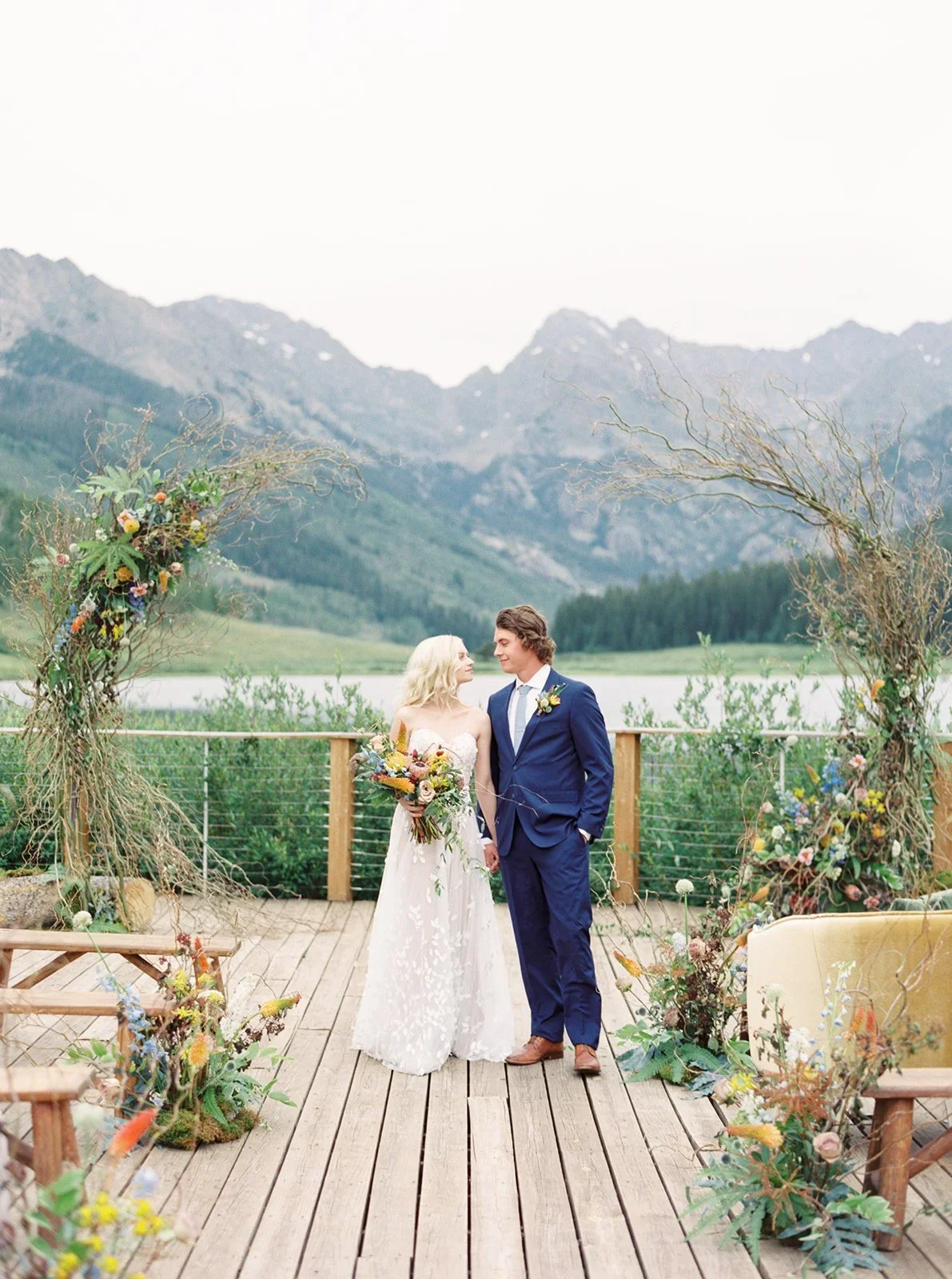 Couple at an outdoor wedding ceremony on a wooden deck with mountain view, decorated with floral arrangements and a rustic arch.