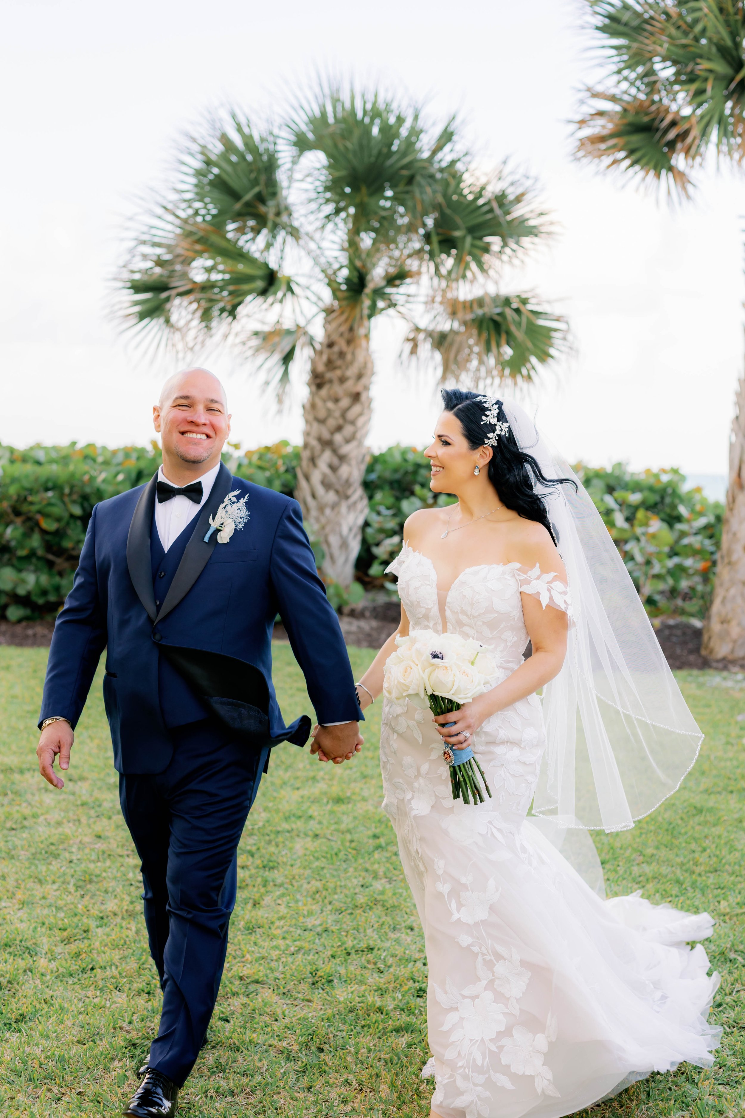 A bride and groom walk hand in hand on grass, smiling, with palm trees in the background. The bride wears a white gown and veil, holding a bouquet, while the groom is in a navy suit.