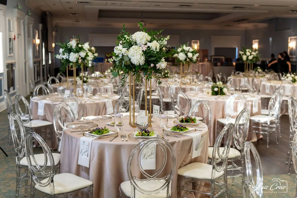 Elegant ballroom setup with round tables, covered in beige tablecloths, decorated with tall floral centerpieces featuring white flowers. Transparent chairs encircle the tables, which are set with plates and salads.