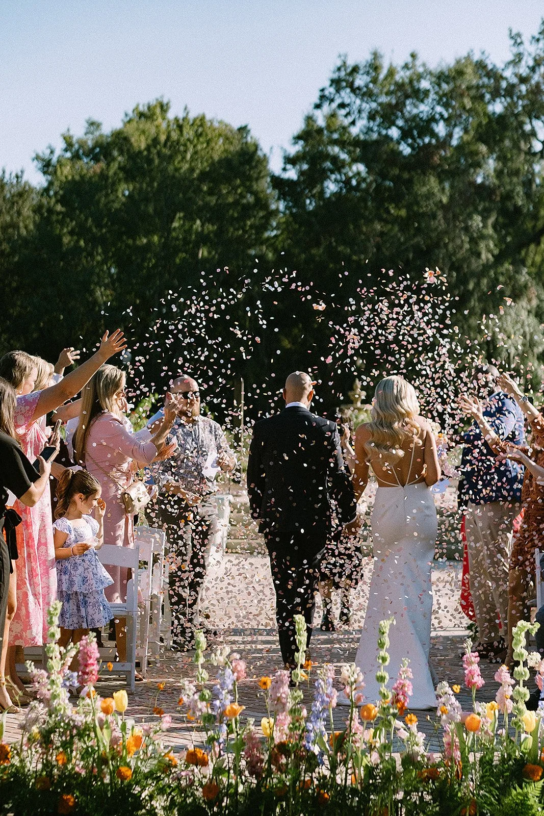 Wedding ceremony outdoors with a bride in a white dress and a groom walking down the aisle while guests throw flower petals. Vibrant flowers in the foreground and trees in the background.