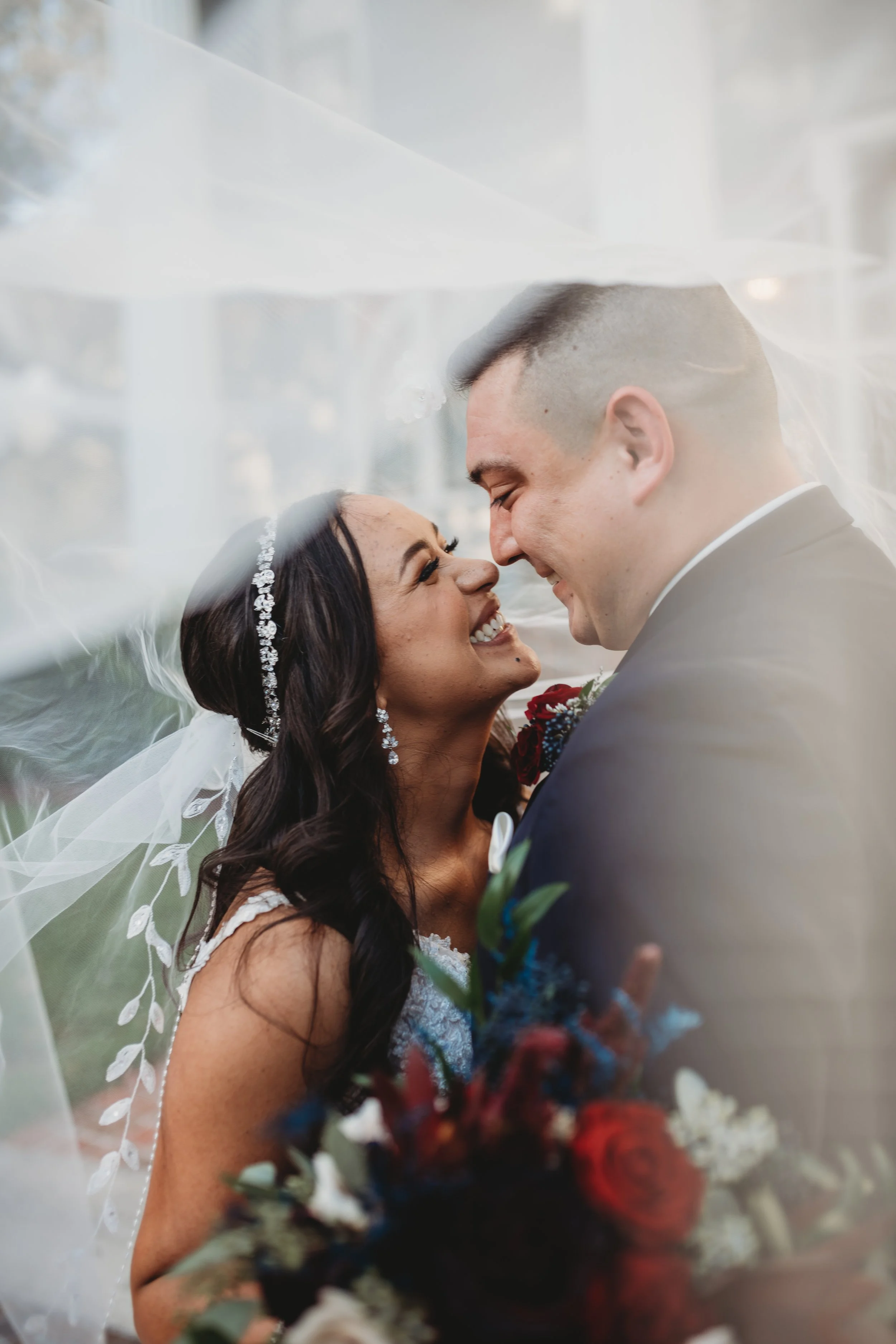 Bride and groom smiling under a wedding veil, holding a bouquet with red and blue flowers.