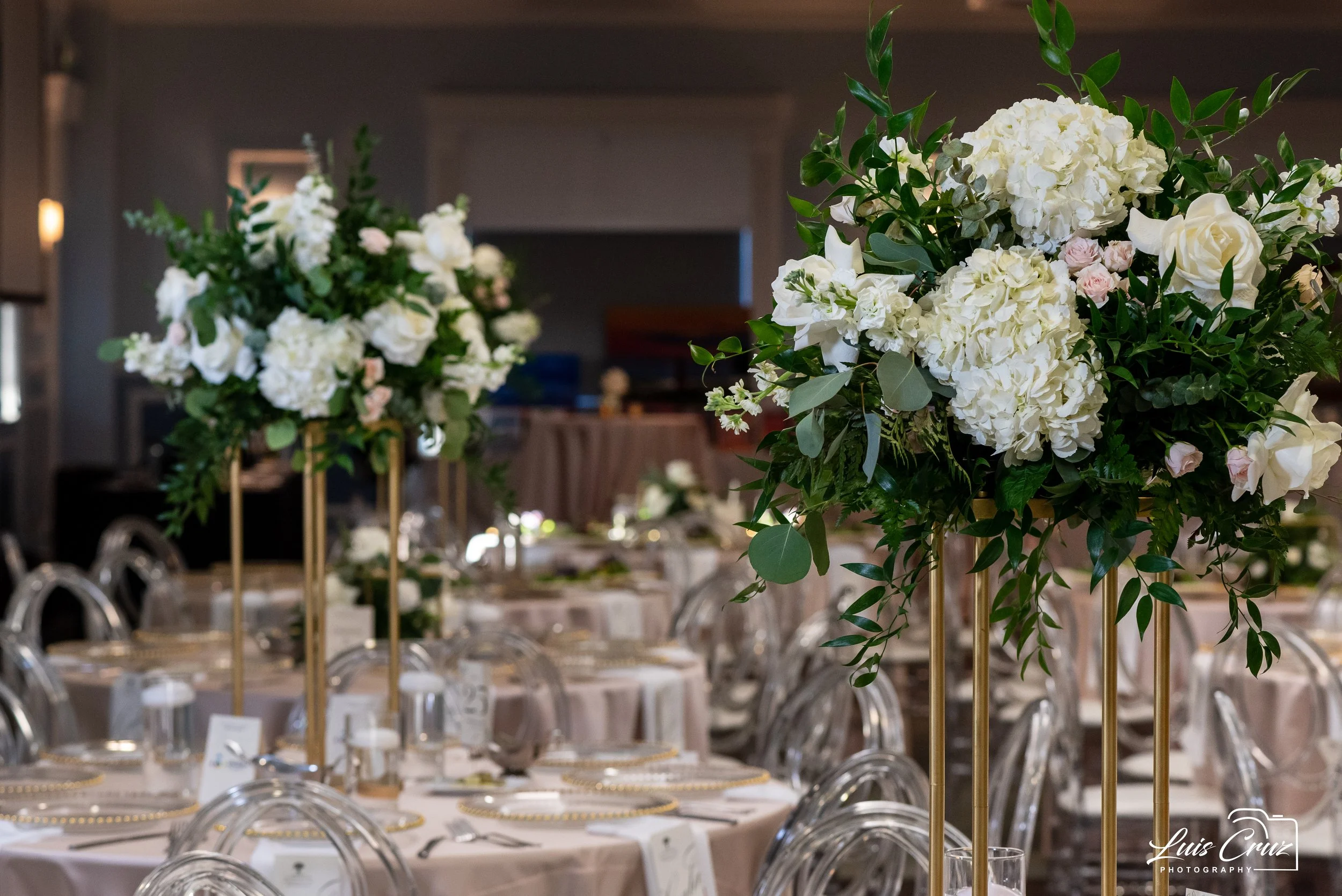 Elegant wedding reception setup with tall floral centerpieces, featuring white roses and hydrangeas, on tables with clear chairs and place settings.