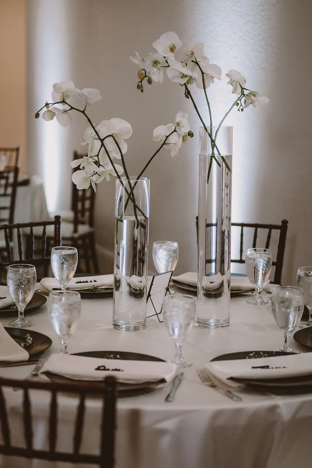 Elegant table setting with white orchid centerpieces in tall vases, surrounded by wine glasses and white napkins on a round table with dark chairs.