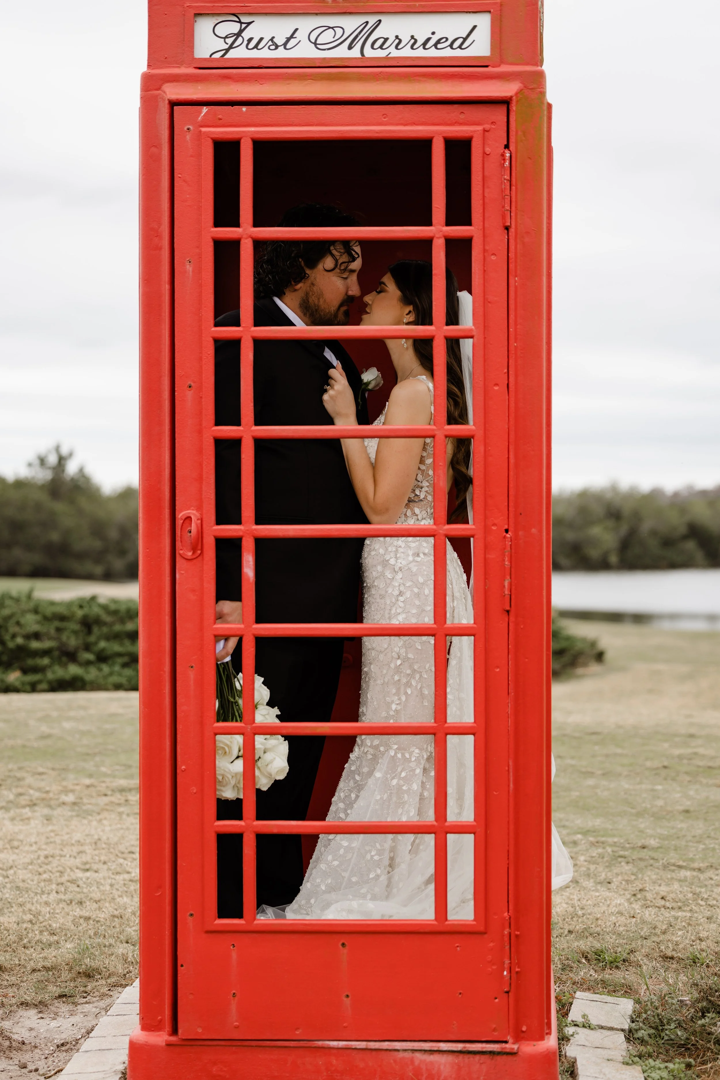 A newlywed couple, in wedding attire, embracing inside a red phone booth inscribed with 'Just Married.'