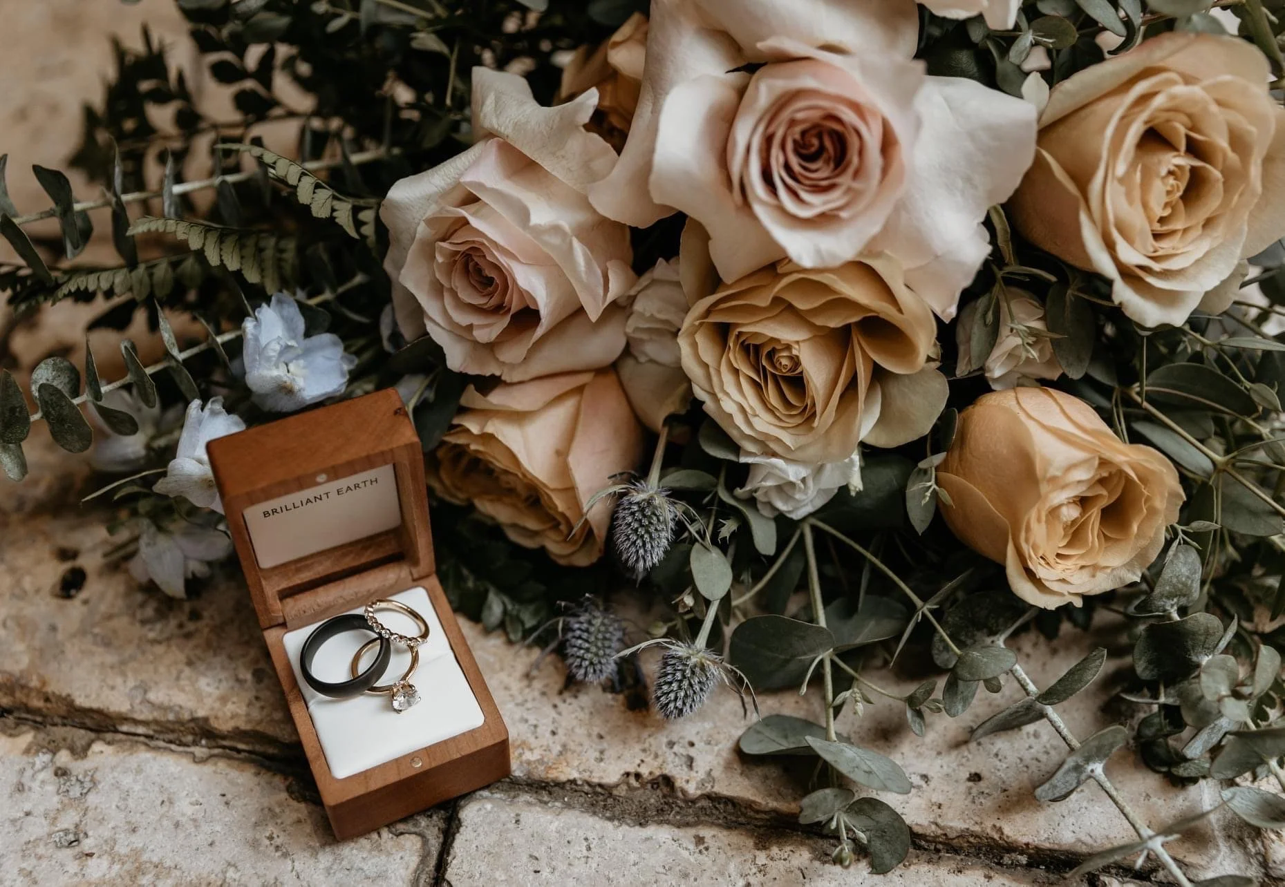 Engagement rings in a wooden box next to a bouquet of cream roses and greenery on a stone surface.