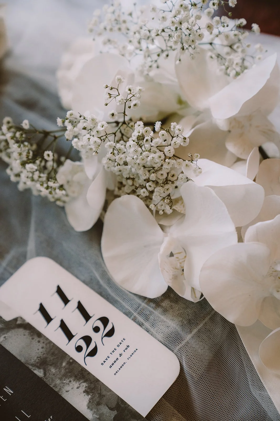 Close-up of white flowers, including baby's breath and orchids, alongside a wedding save-the-date card with "11 11 22" displayed on it.