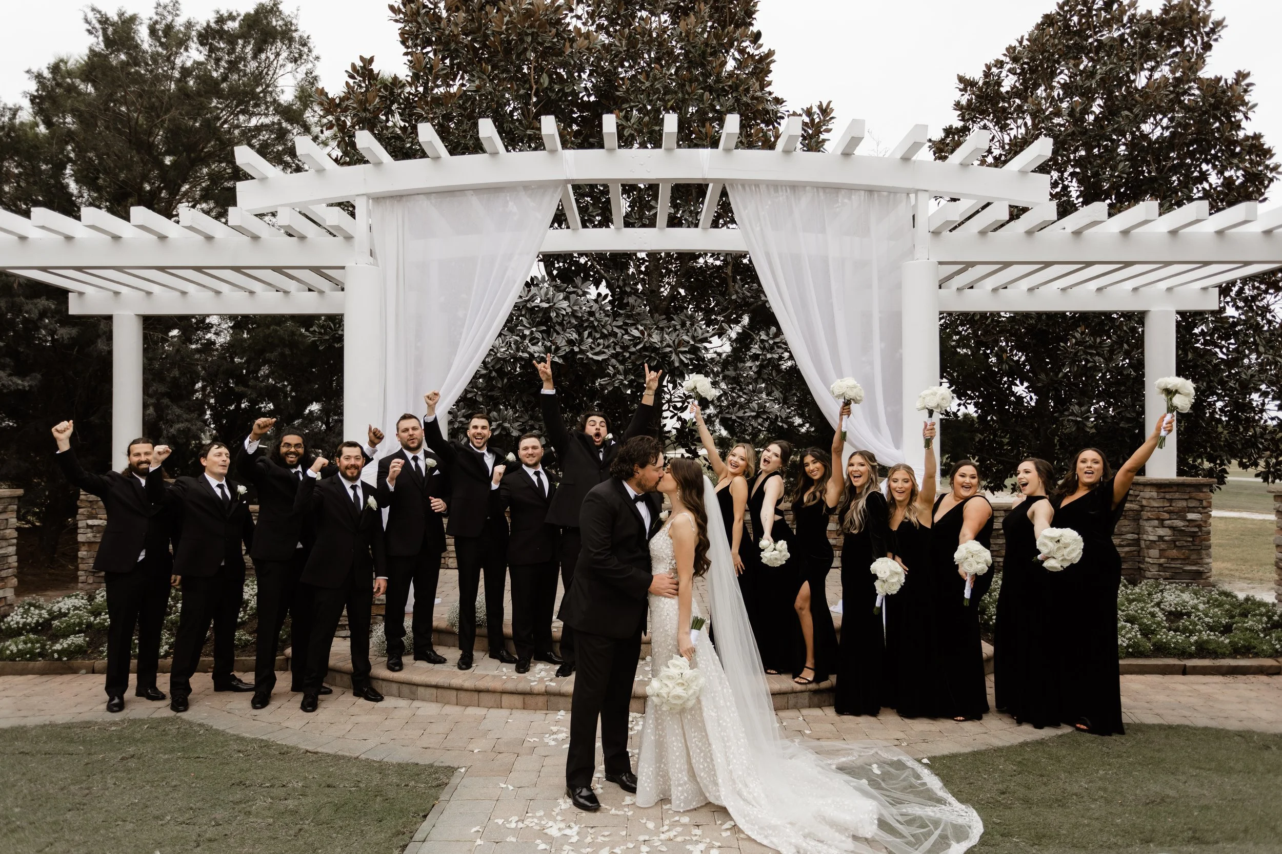 Bride and groom kissing under a white pergola, surrounded by a cheering bridal party wearing black attire, holding bouquets, outdoors.
